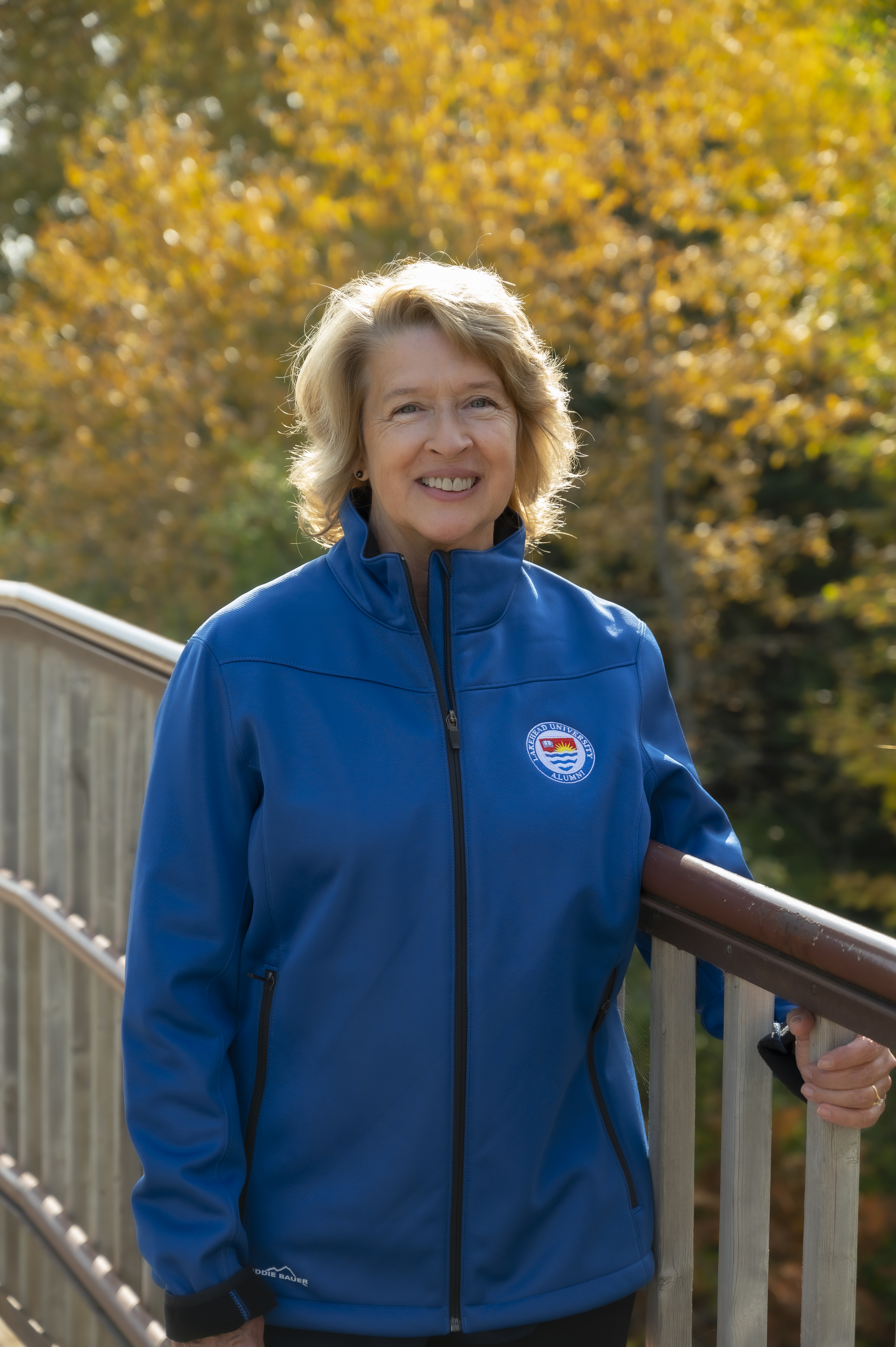 Dr. Moira McPherson standing on foot bridge in the autumn wearing a Lakehead alumni jacket
