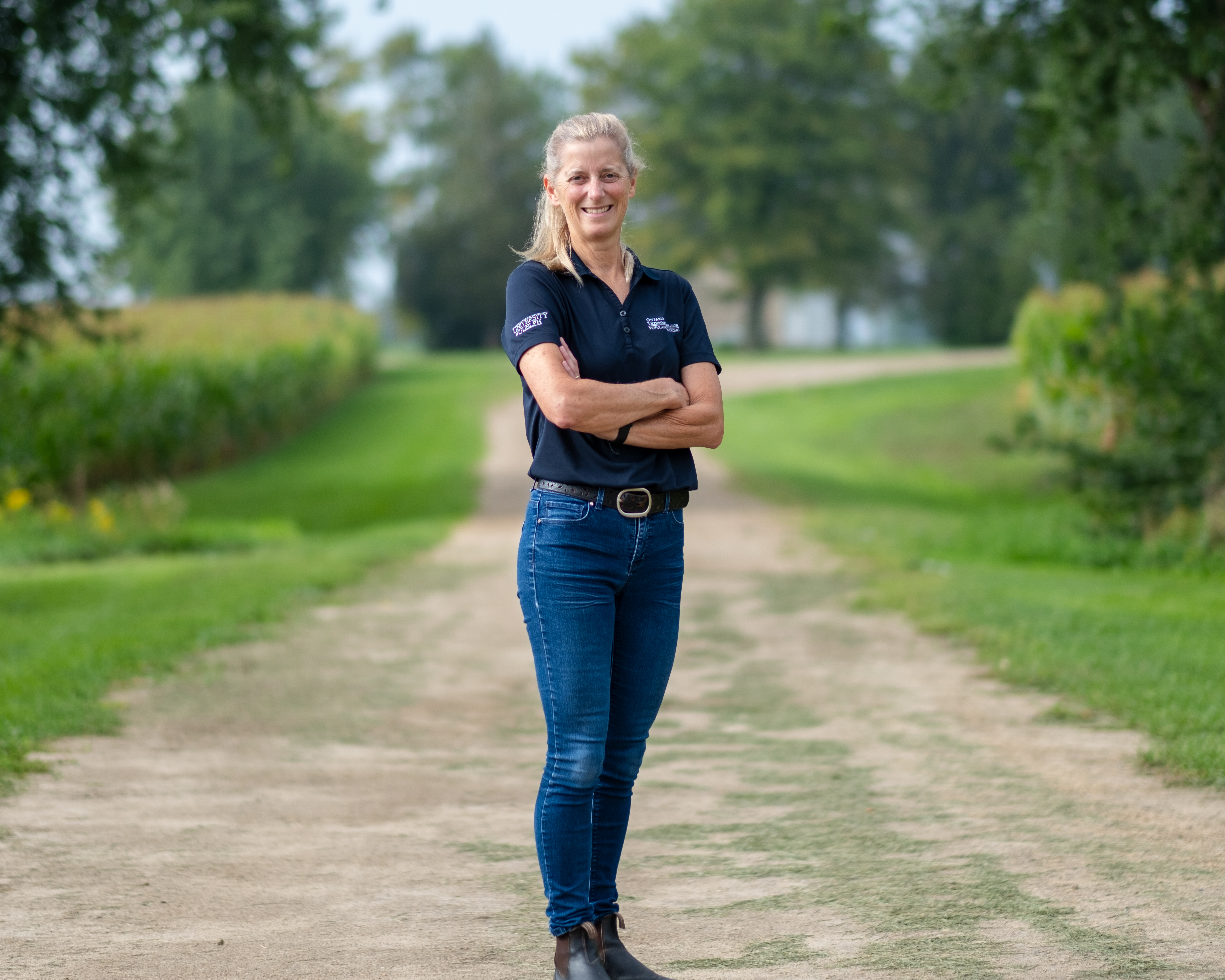 Woman standing on a dirt road flanked by grass and trees and crops. She is wearing jeans and a golf collared t-shirt and has her arms crossed smiling at the camera.