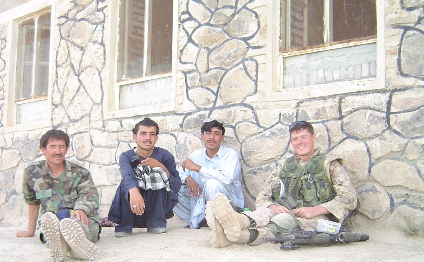 Robin Rickard dressed in combat gear sits outside a building in Afghanistan with three other men.