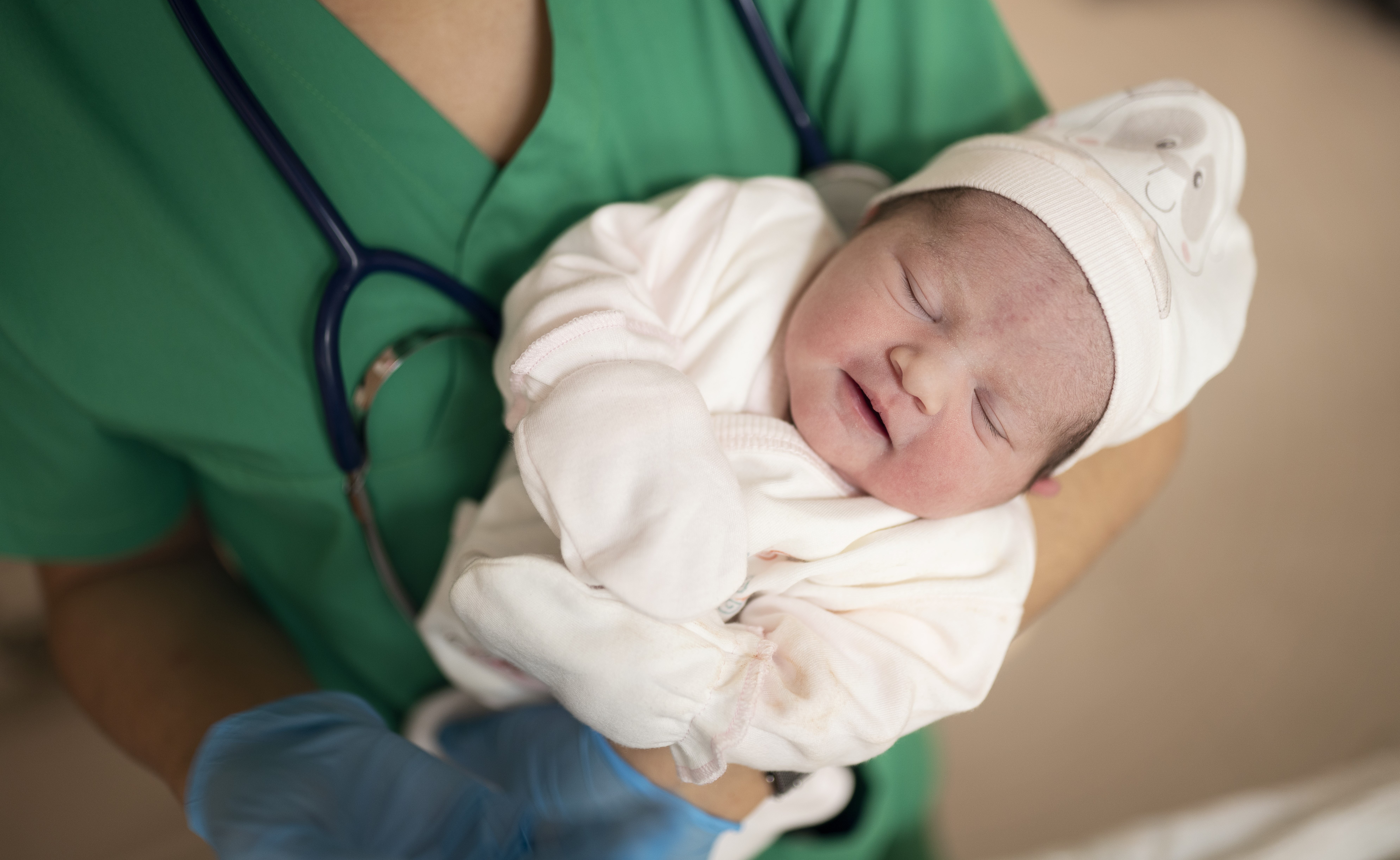 A doctor wearing green scrubs with a stethoscope around her neck holds an infant wearing white footie pajamas and white cap