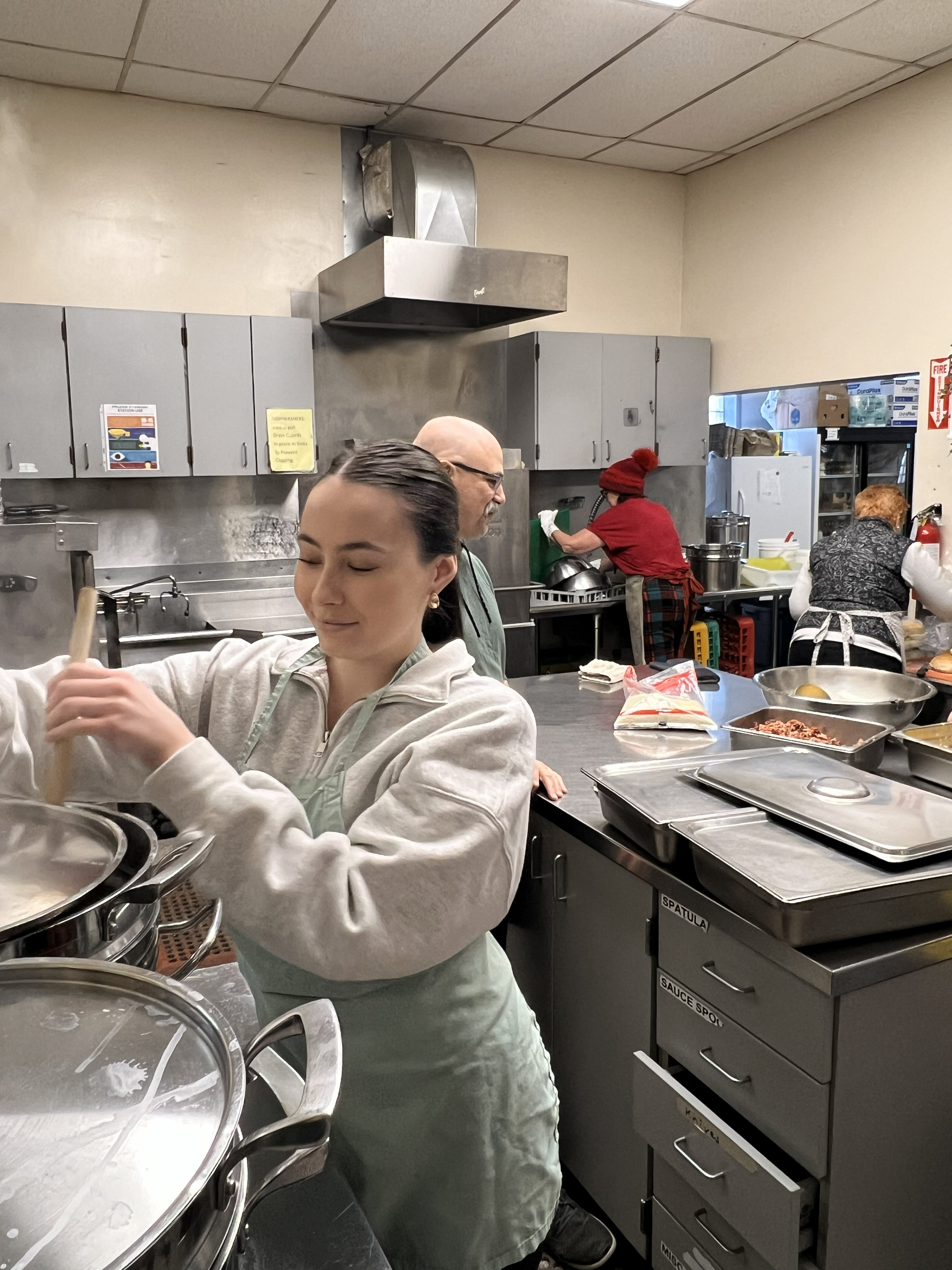 Bianca Zussino stirs a large pot on a stovetop while other Dew Drop Inn volunteers help prepare food in other parts of the kitchen