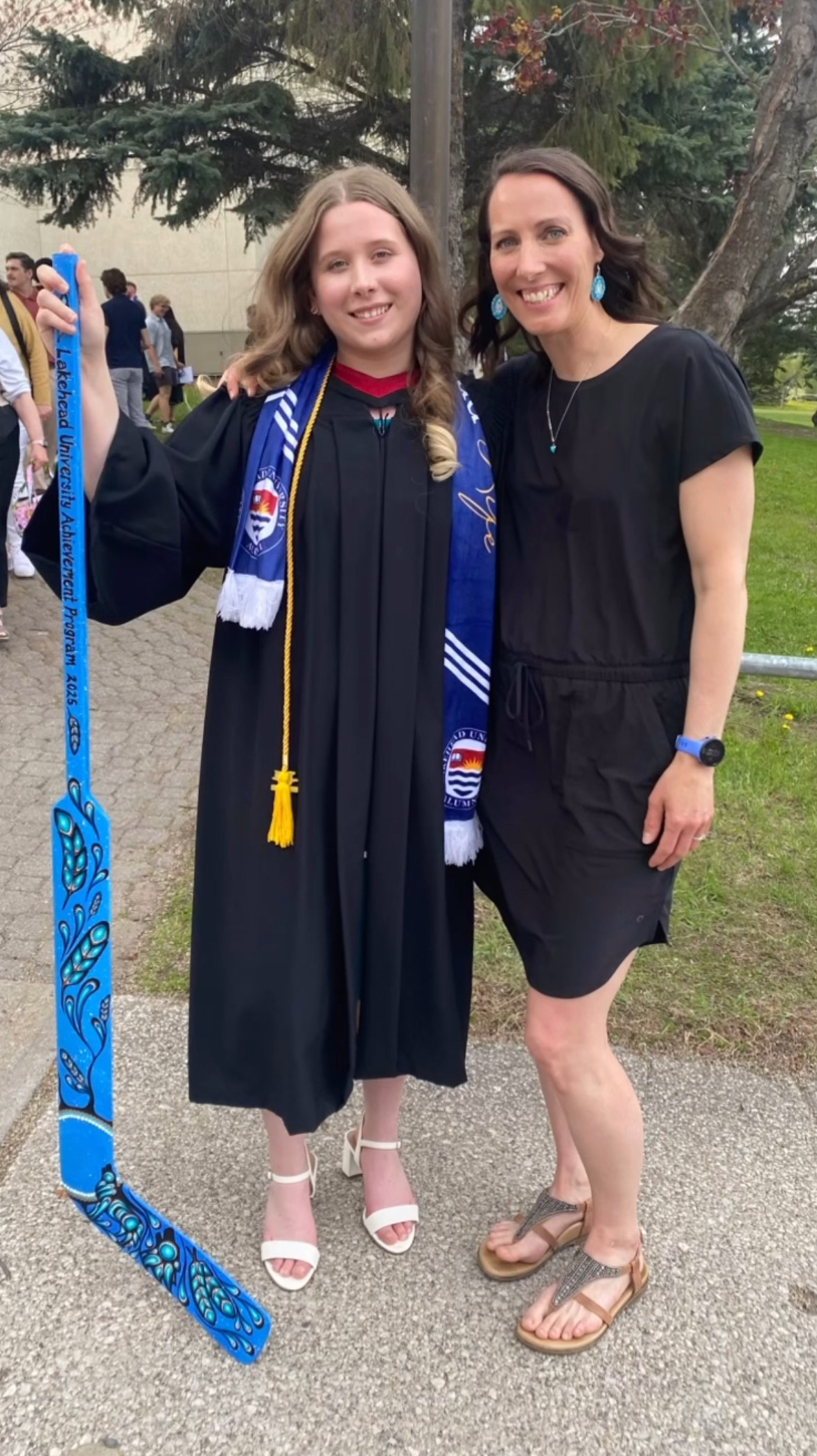 Camryn Williams wearing graduation robes and holding an Achievement Program hockey stick stands beside Achievement Program Coordinator Amanda Stefanile after a 2025 Lakehead convocation ceremony