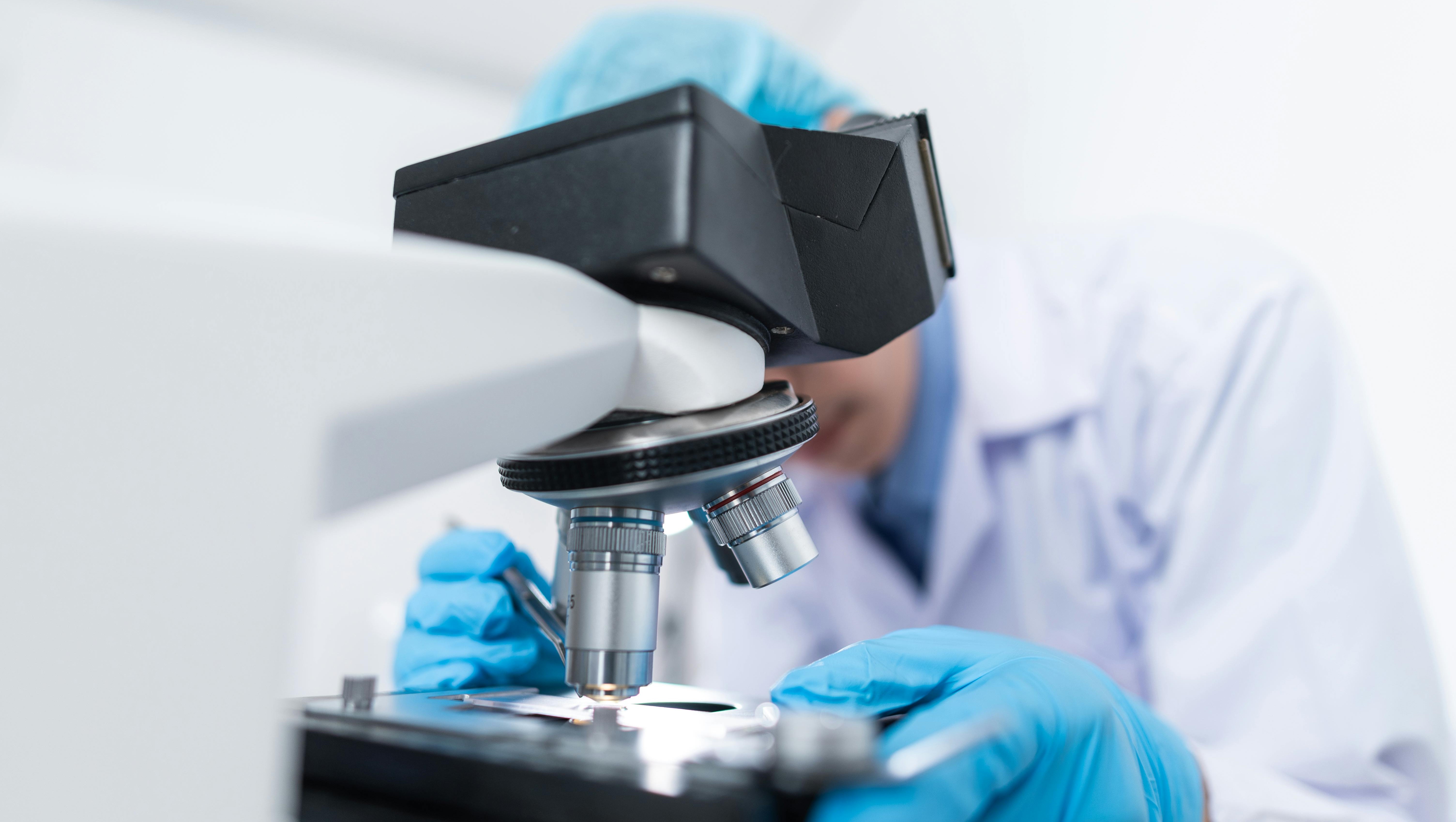 Lab Technician Using a Microscope A lab technician wearing gloves and a lab coat uses a microscope