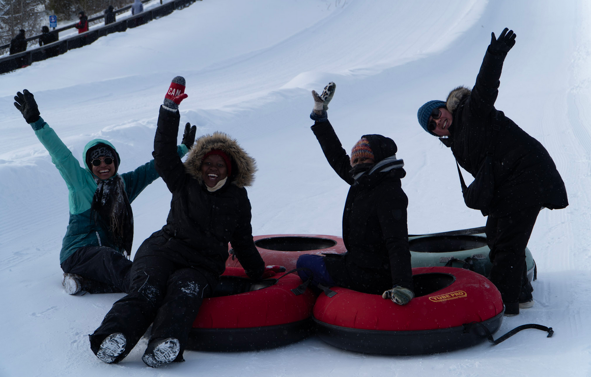 Students Tubing Four smiling students on inner tubes at the bottom of a snow-covered ski hill each wave a raised hand in the air