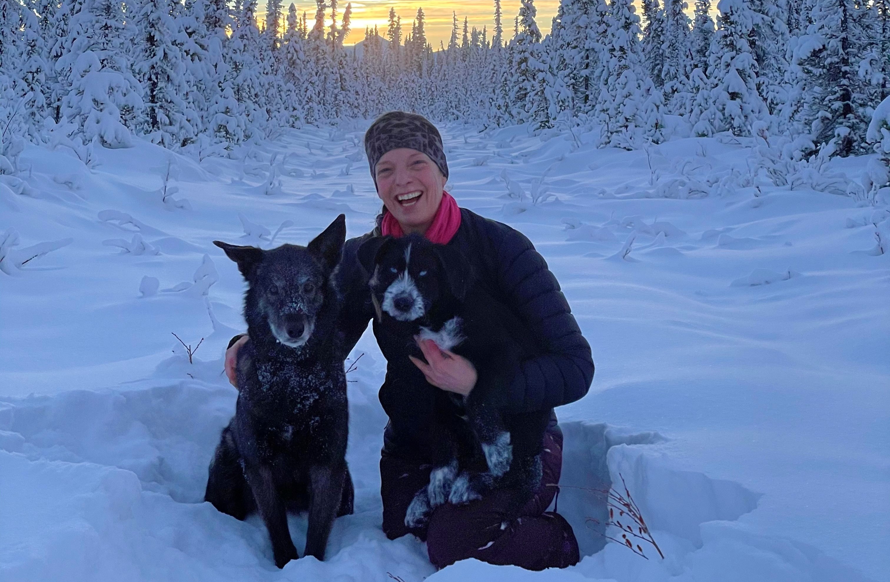 Heidi Warren sits in the snow, with snow-covered trees behind her, while hugging two dogs 