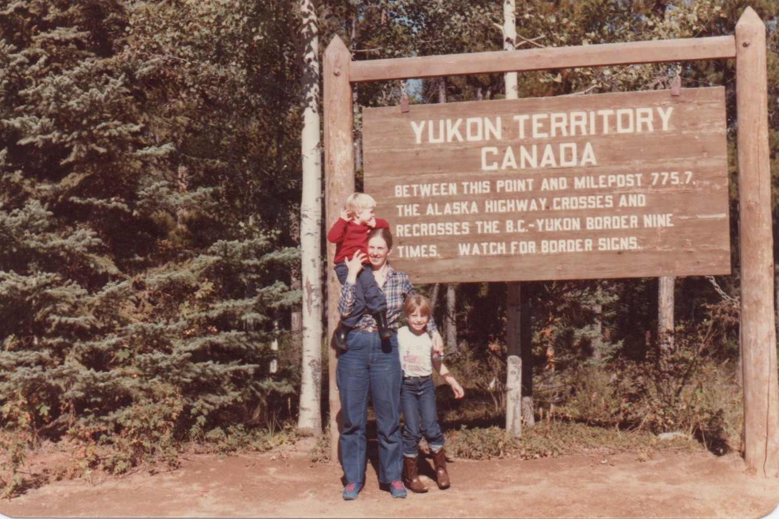 Heidi Warren's mother stands in front of a sign saying Yukon Territory Canada; her arm is around Heidi and she holds Heidi's sister on her shoulder 
