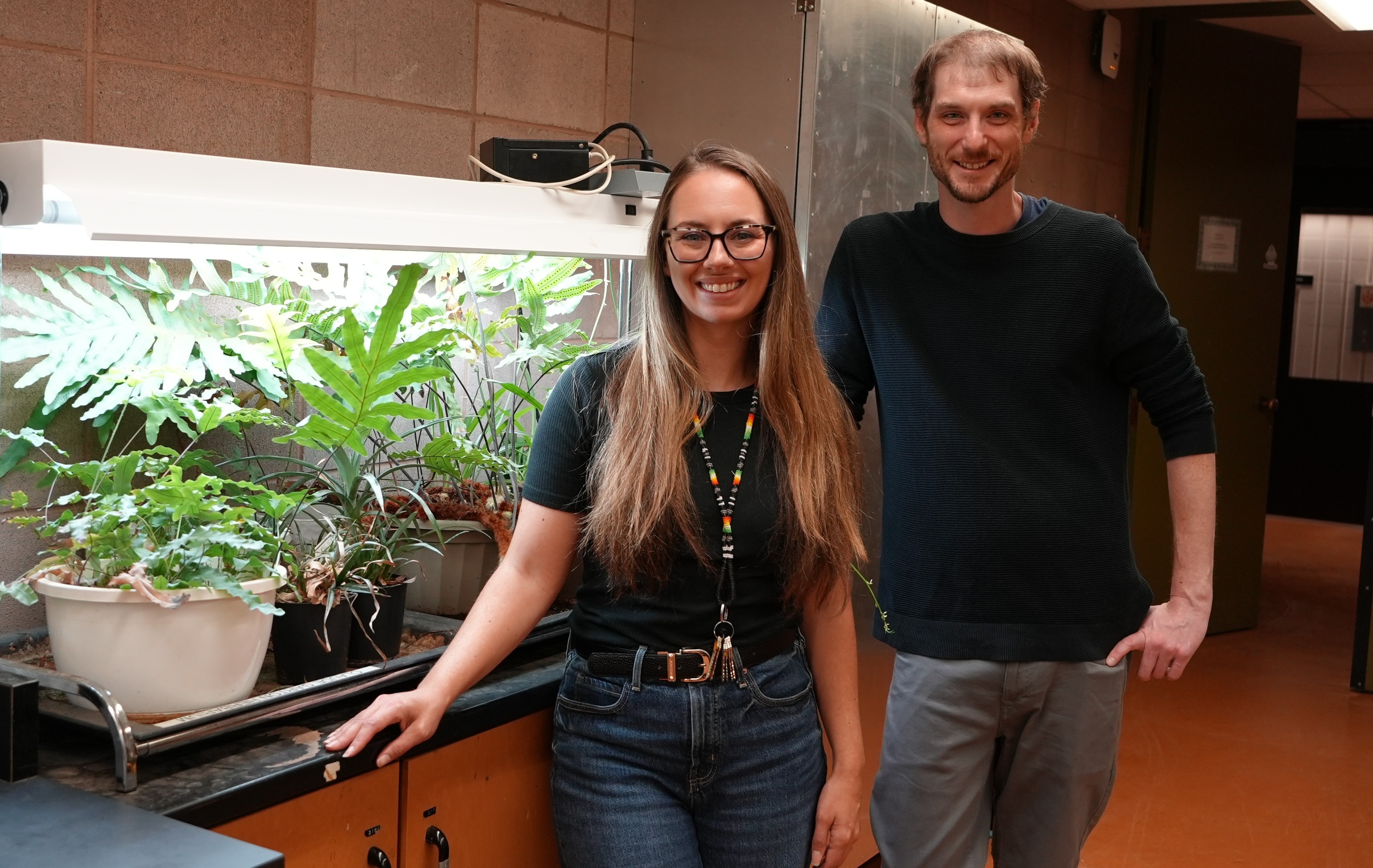 Kristi Valley and Myles Cummins stand in front of potted plants on a countertop near the entrance of the Claude E. Garton Herbarium