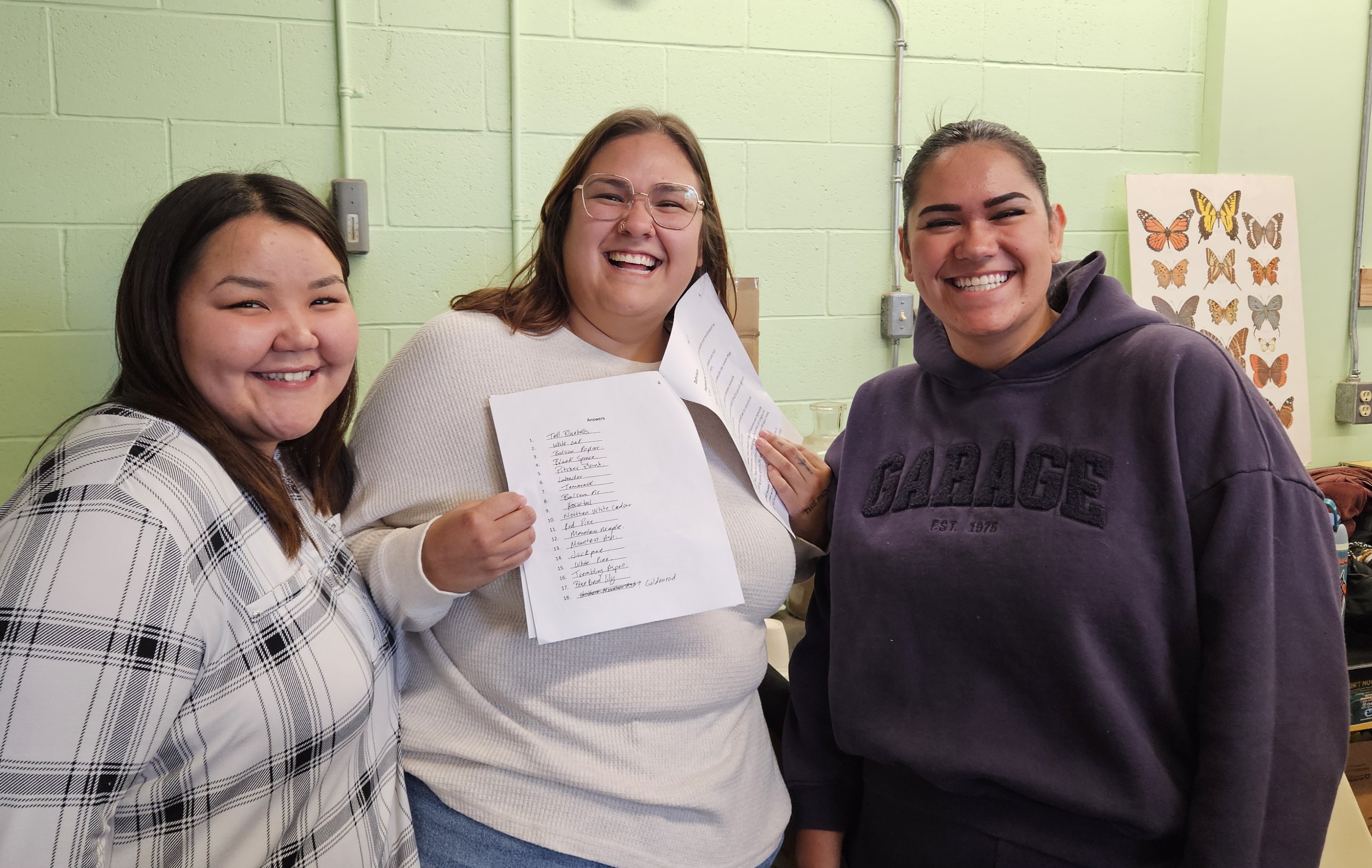 Three students standing in a classroom; the student in the centre holds up a list of plant and tree names