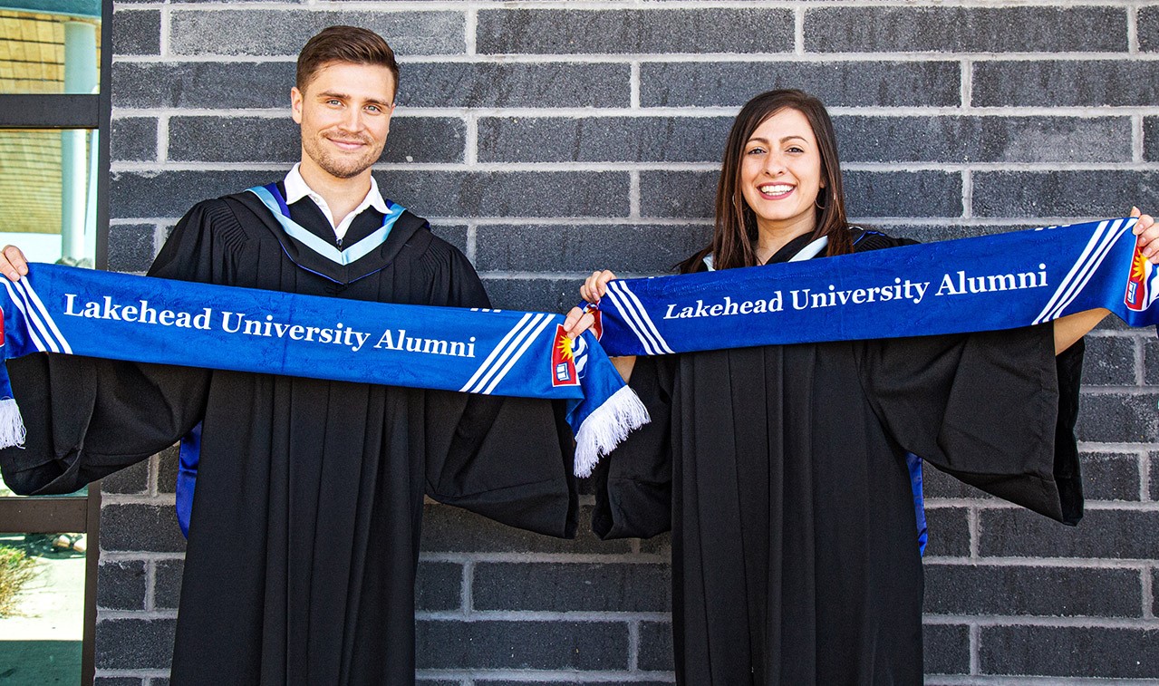 A male graduate and a female Lakehead graduate wearing convocation robes stand outside on the Orillia campus holding Lakehead alumni scarves