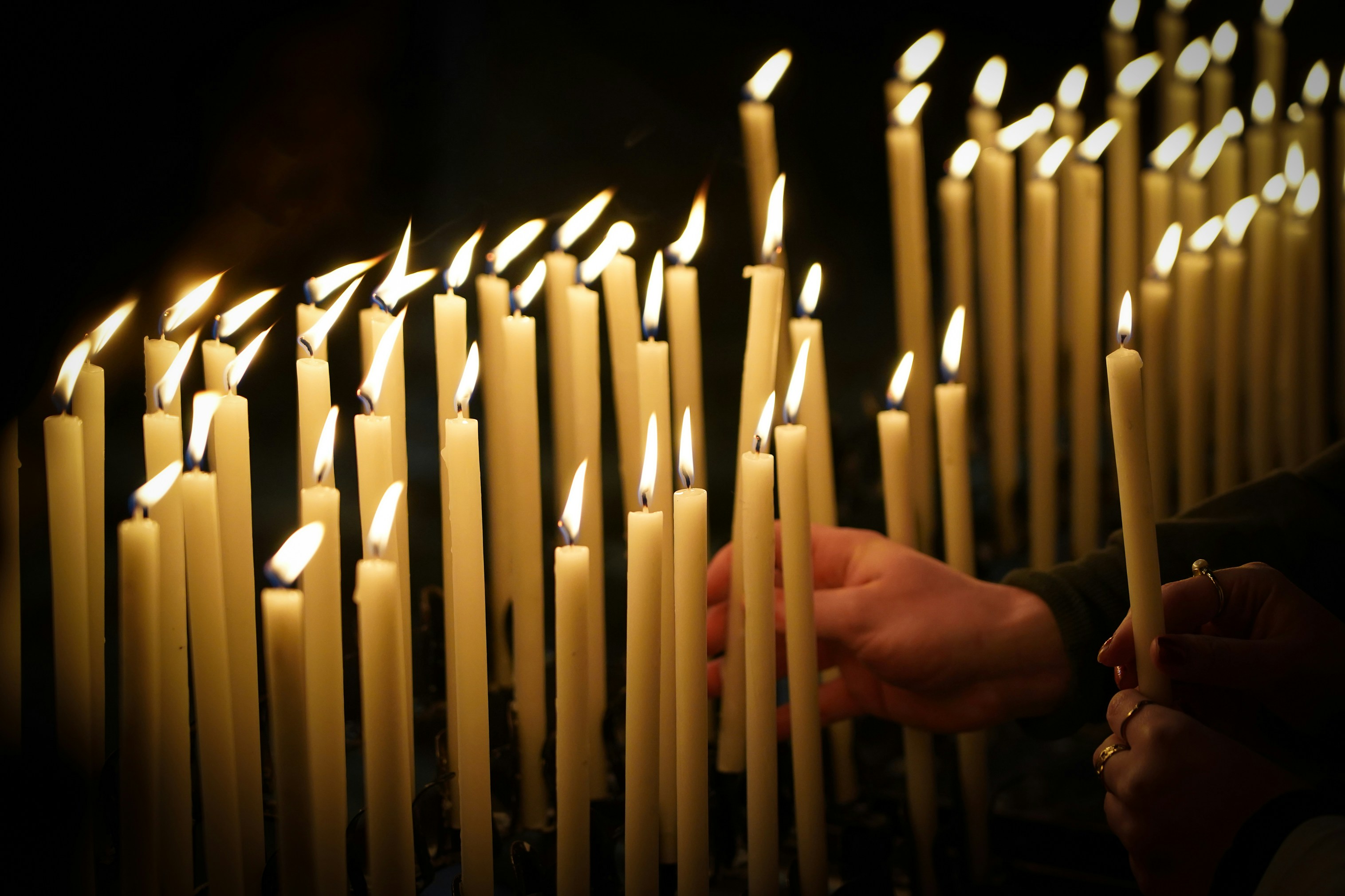 The hands of two people holding lit candles add their candles to a group of white candles burning in the darkness