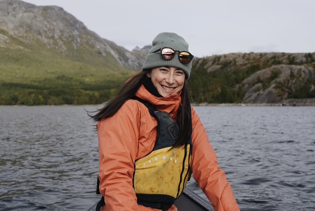 Teigan Labor, wearing a waterproof jacket and a life jacket, sits in a canoe on the water with rocky mountains in the background