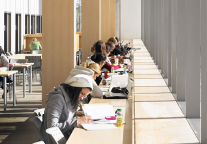 Several students sitting in a row of desks facing a large window. Books, water bottles and cups covers the desks.