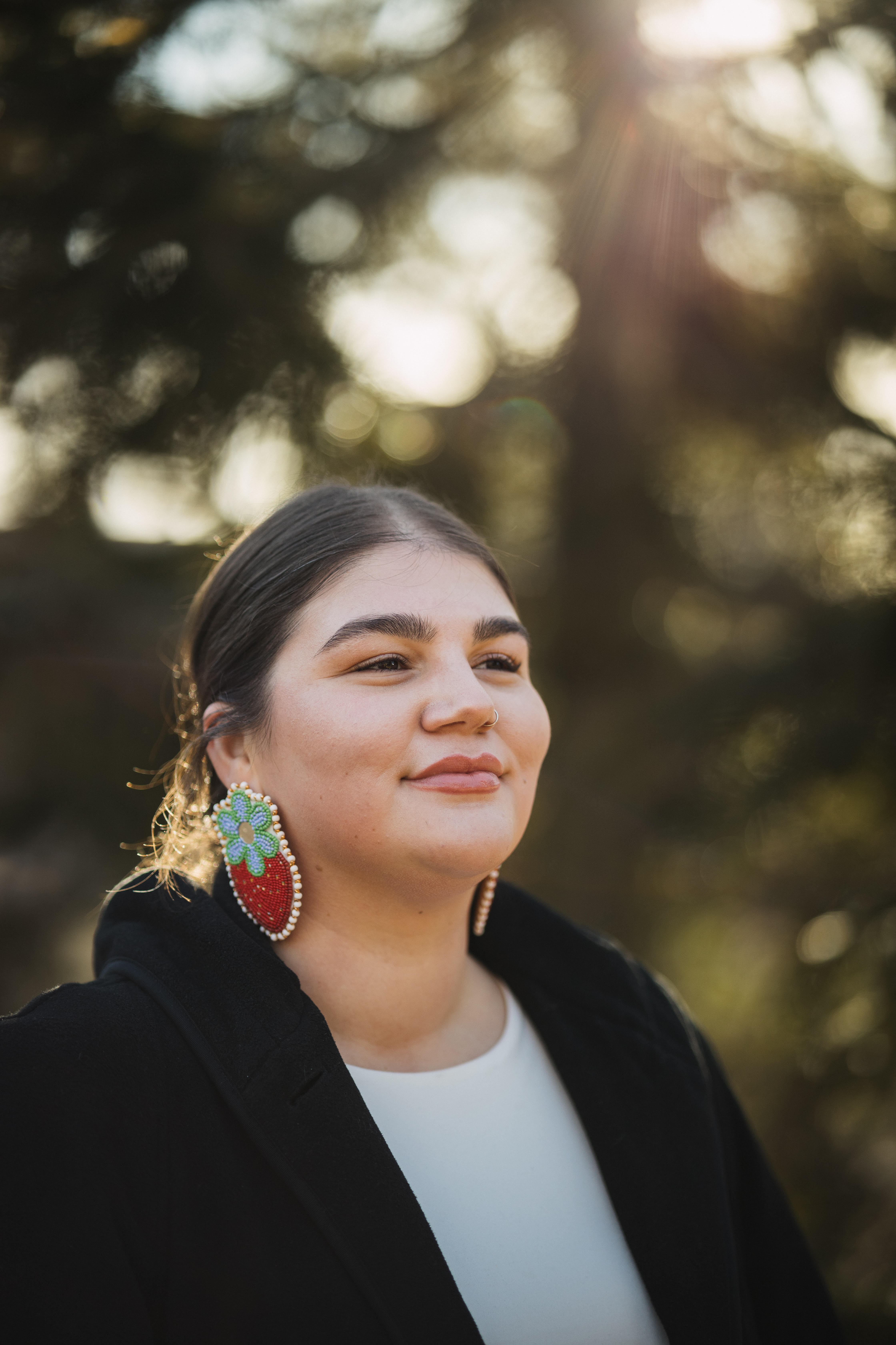 A woman wearing a black coat over a white shirt. She is wearing beaded earings and her brown hair is tied back.
