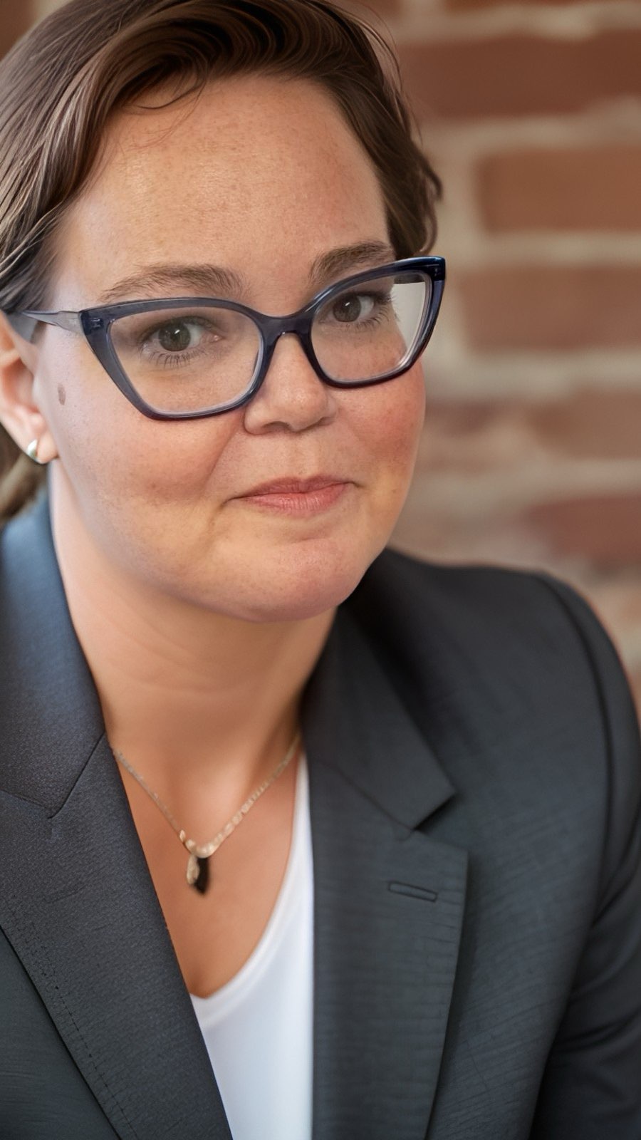 Woman wearing glasses with brown hair tied back and wearing a suit.