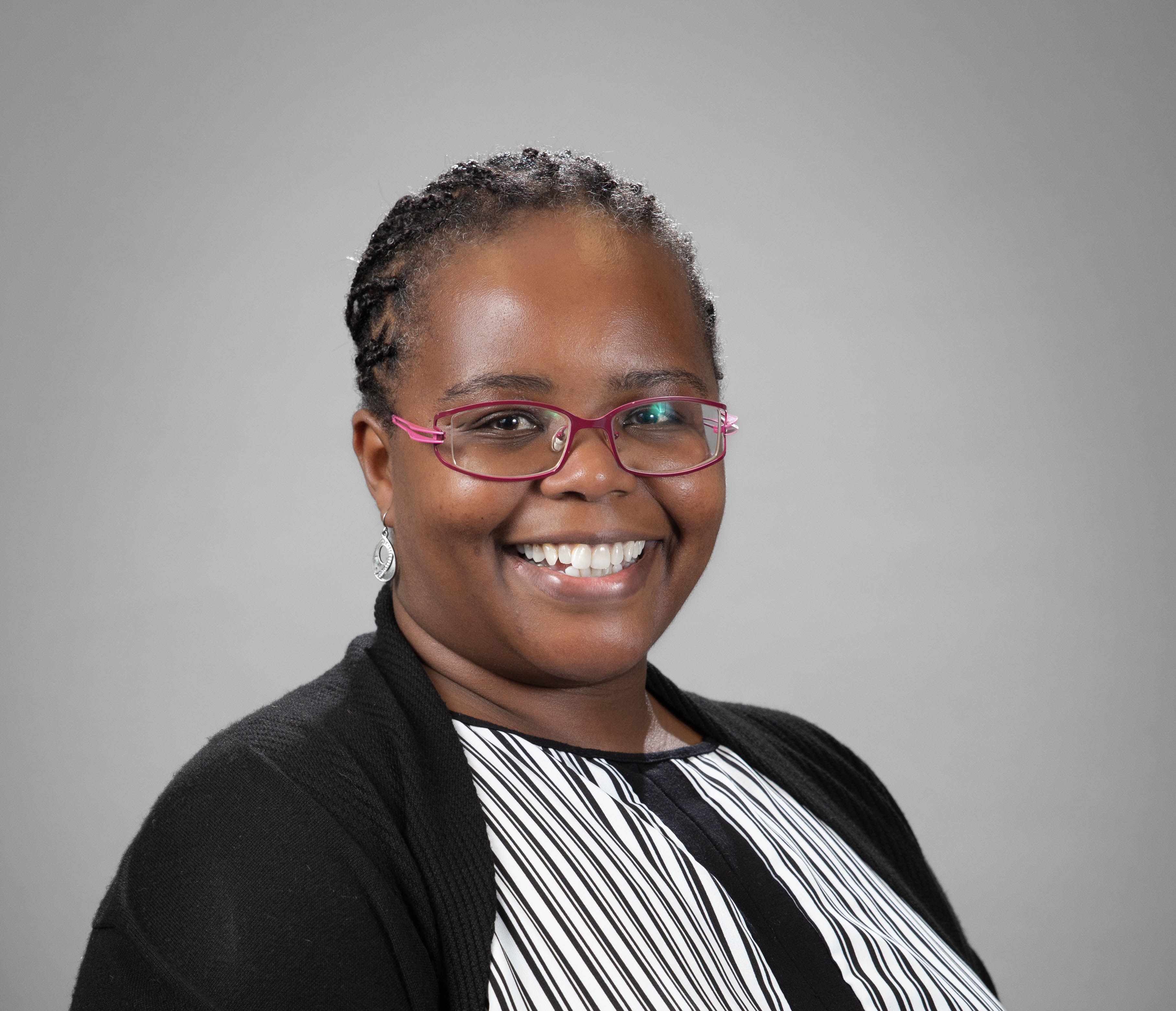 A smiling woman wearing glasses with a black and white stripped dressshirt and a black cardigan. She is Black and the picture has a grey background.