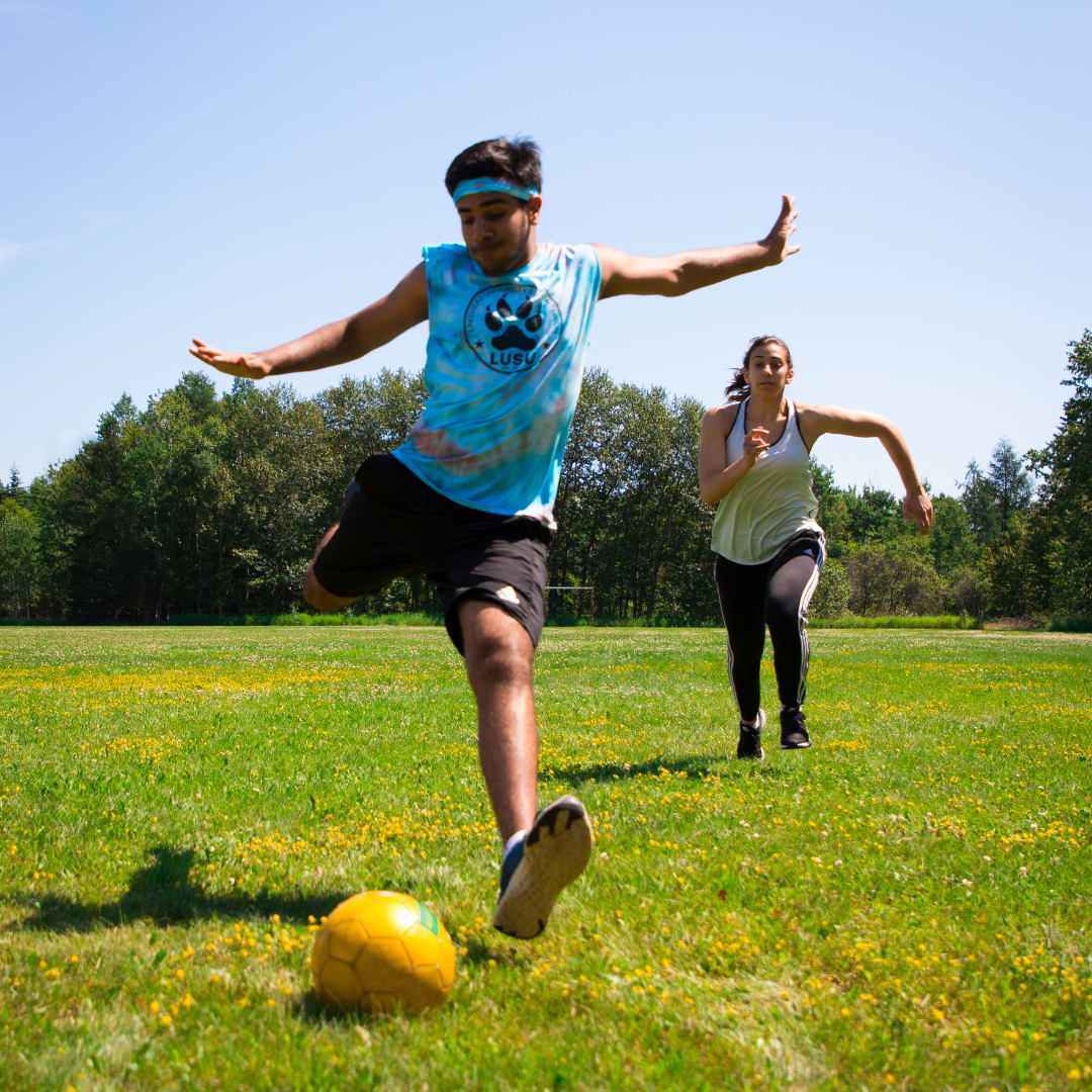 Person with outstretched arms winding up to kick a yellow soccer ball on a grass field. Person chasing them in the background,