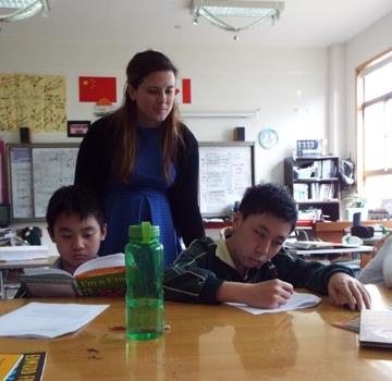 This is a picture of a teacher who is working with students. One student is reading a book and the other is writing
