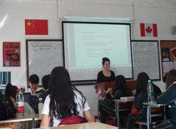 This is a picture of a teacher giving a lesson to a class. She is standing at a lectern while the students are sitting at their desk