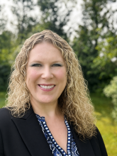 Ashleigh is a caucasian female with blonde curly mid-length hair and is pictured wearing a black suit jacket standing in front of a background of green trees