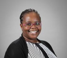 A smiling woman wearing glasses with a black and white stripped dressshirt and a black cardigan. She is Black and the picture has a grey background.