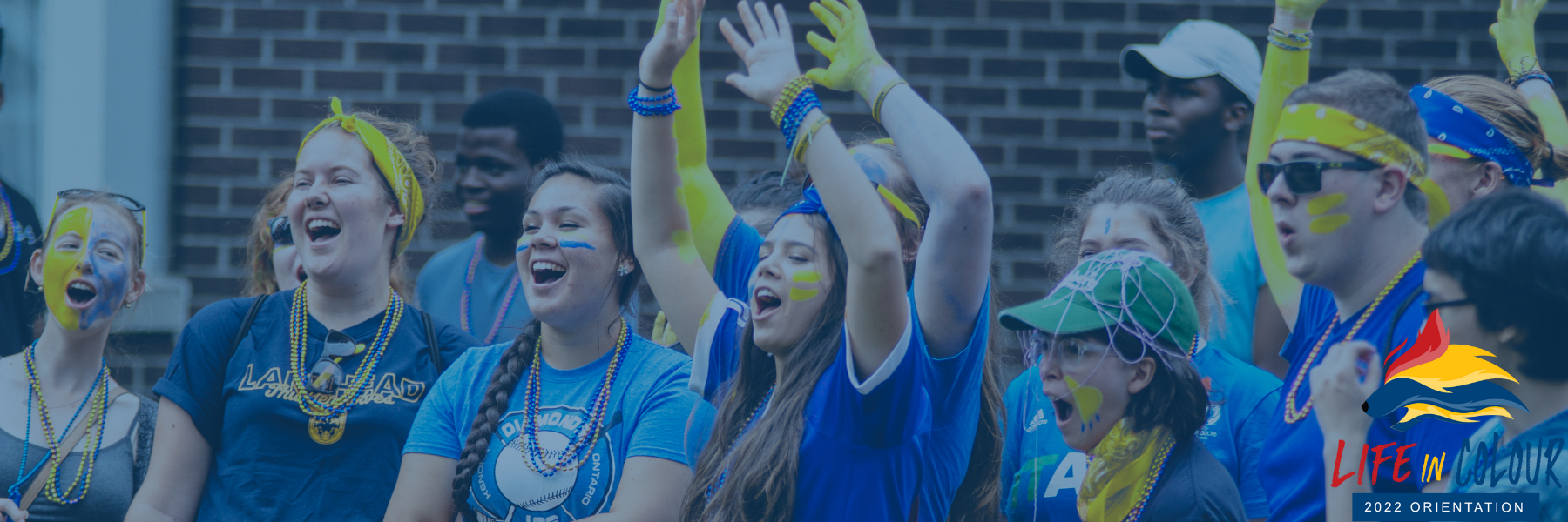 Group of Lakehead students wearing blue and yellow spirit wear cheering and smiling while having fun at Orientation. The Life in Colour, Orientation 2022 rainbow wolf head logo is located in the bottom right corner of the image.