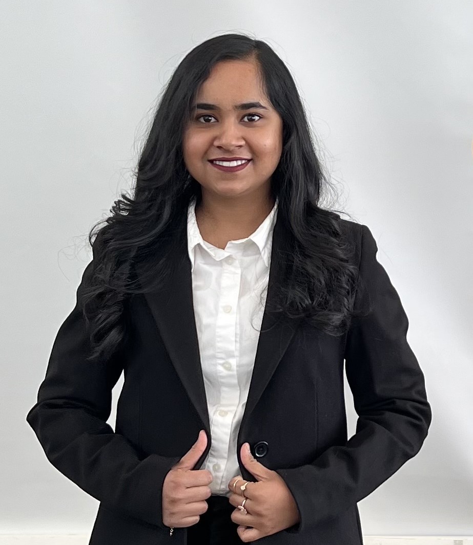 Rechal Parmar Portrait Image Rechal Parmar, wearing a white shirt and black suit, stands in front of a white backdrop