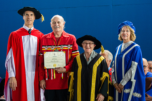 A tall man with a black cap wearing red and white regalia, a man of Anishnaabe heritage, a woman wearing black and yellow regalia, and a woman wearing blue and white regalia with blue cap
