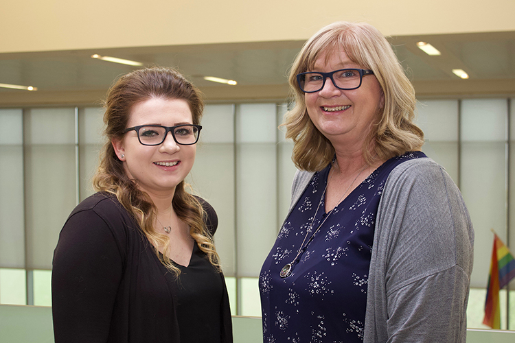 A young woman with brown hair stands beside her mother on a balcony overlooking a green field