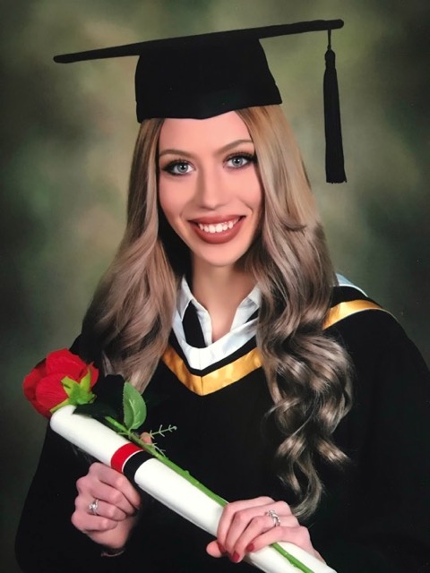A blonde haired women wearing a graduate cap is smiling at the camera