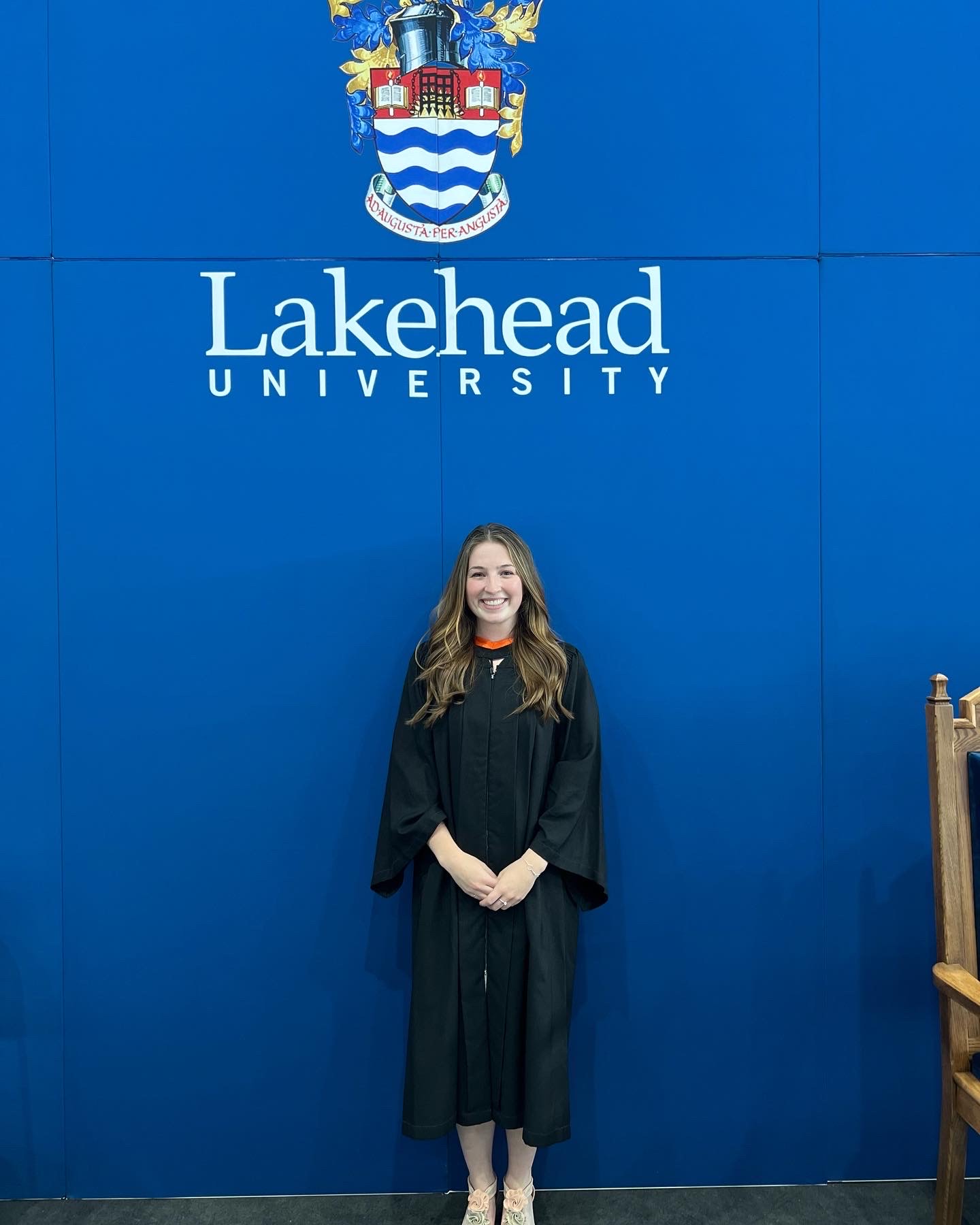 Lakehead student Avery Williams in her graduation robes standing in front of a Lakehead University screen with the school crest