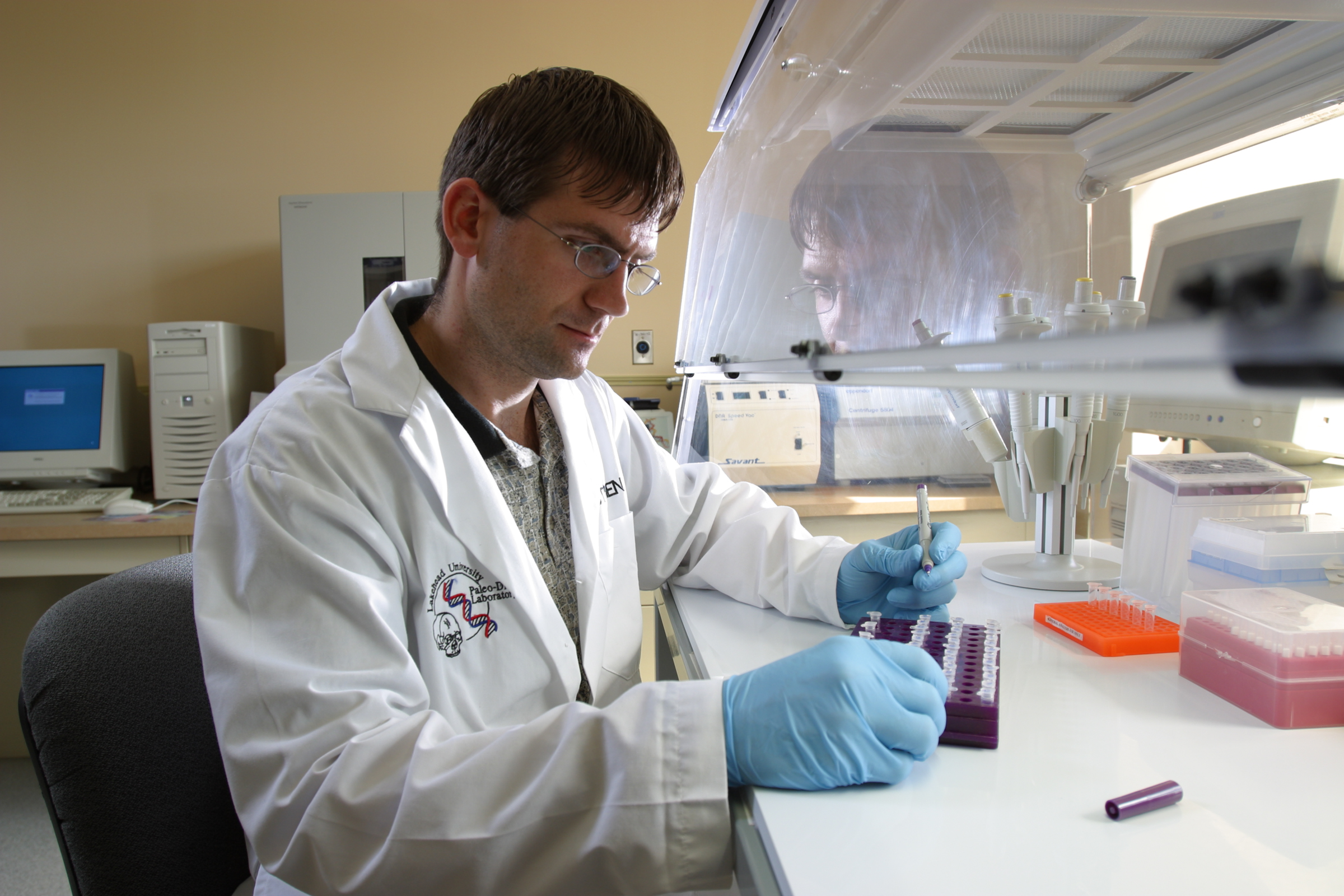 Stephen Fratpietro, wearing a lab coat and gloves, looks at a tray of samples while holding a marker