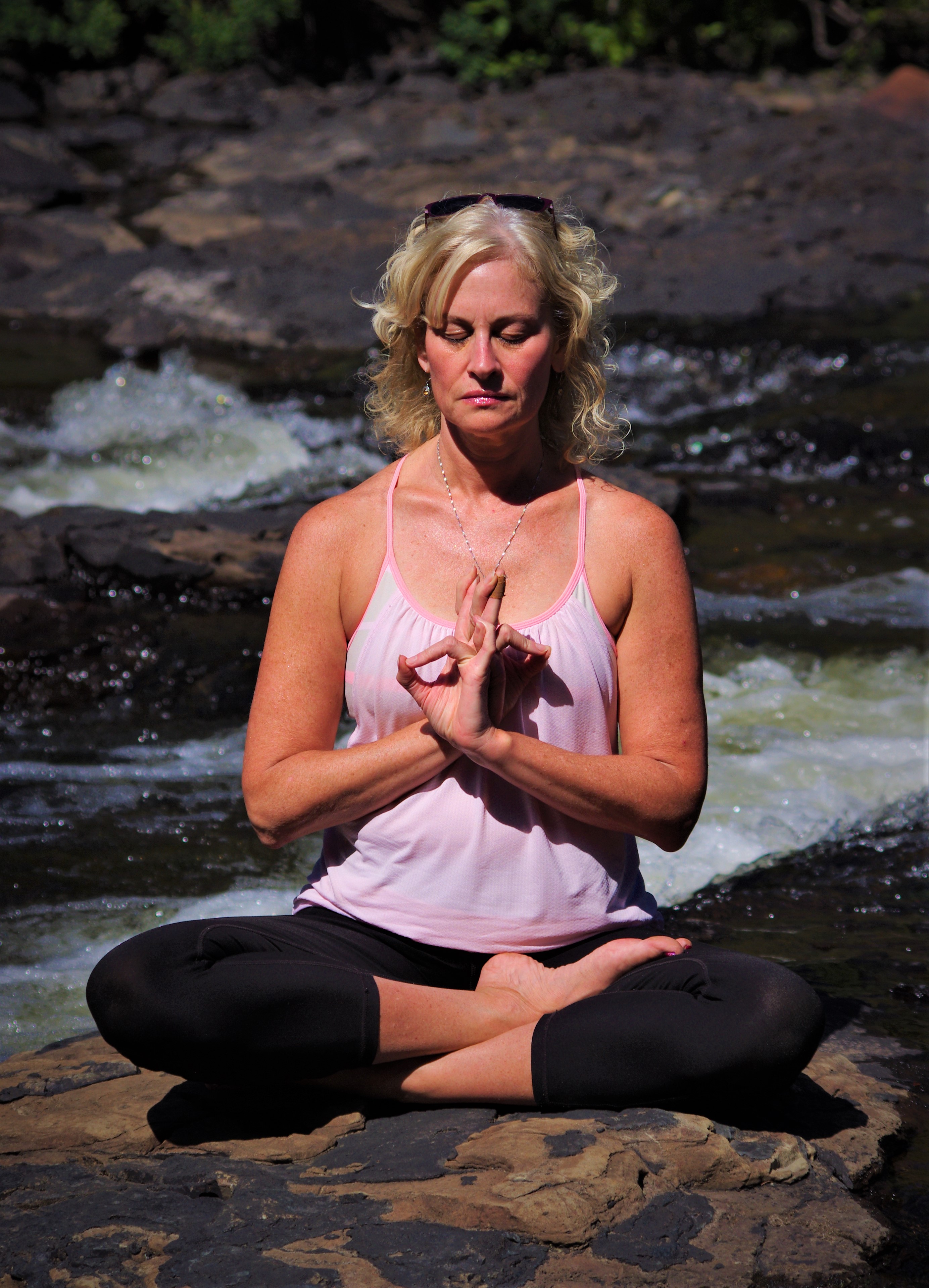 Angela sits cross legged by a river in a yoga pose
