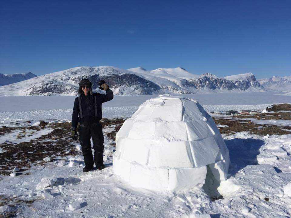 Ian McRae in hat and winter clothes standing beside an igloo waving