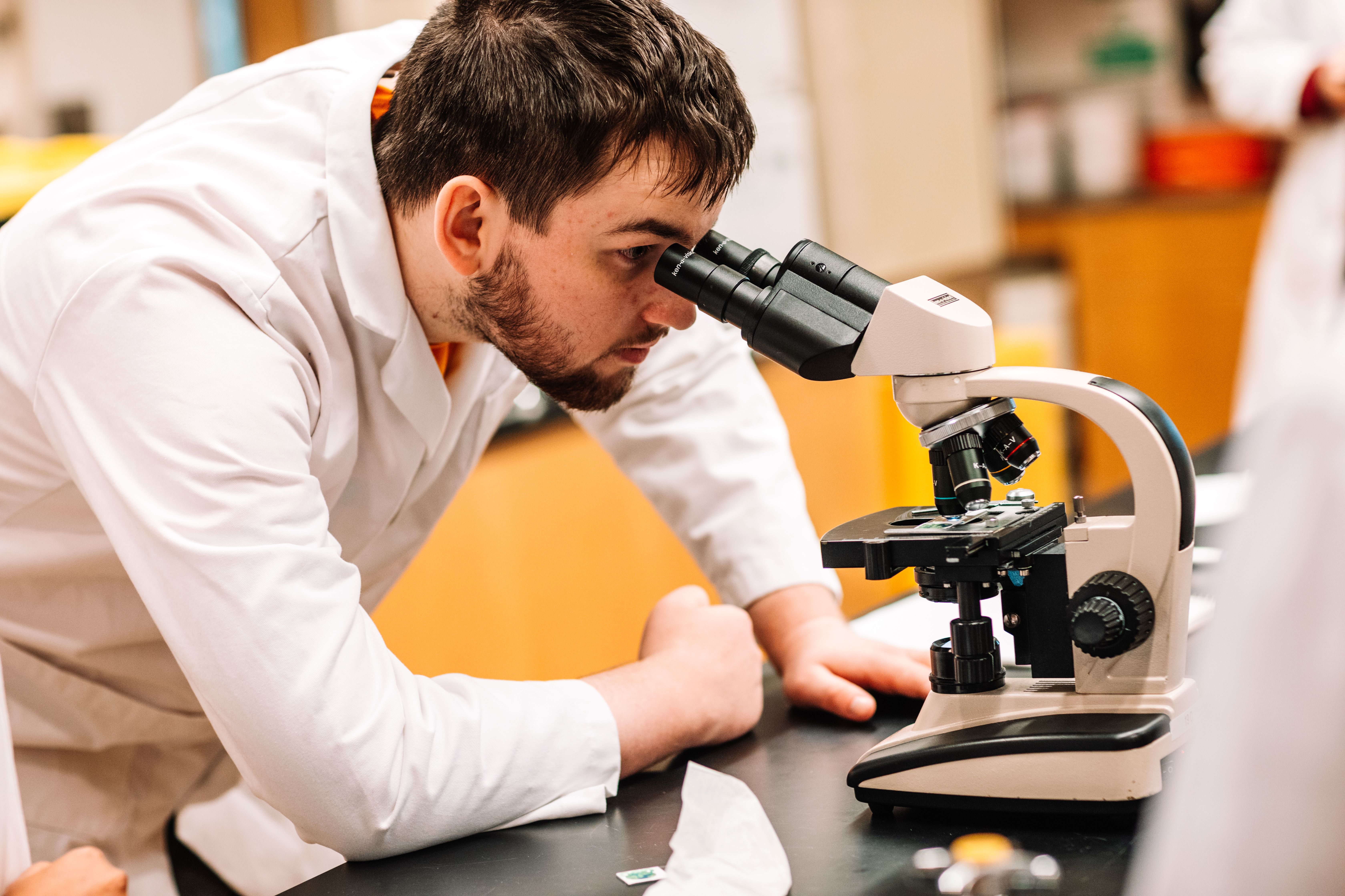 A male Lakehead Orillia student in a lab coat peers into a microscope