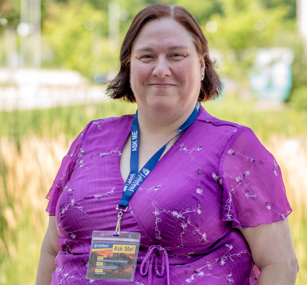 Dr. Meridith Lovell-Johnston wearing a purple shirt and Lakehead Univeristy lanyard stands outside on a sunn day.