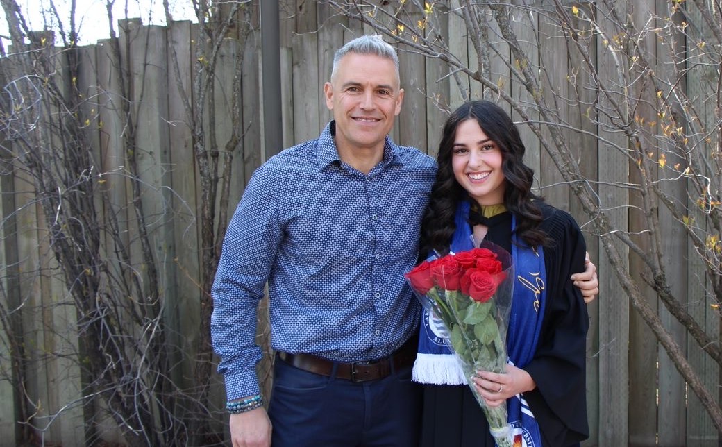 Enzo Fabiano stands with his arm around his daughter Madeline Fabiano who is wearing a black convocation robe and a blue scarf and who is holding a bouquet of red roses