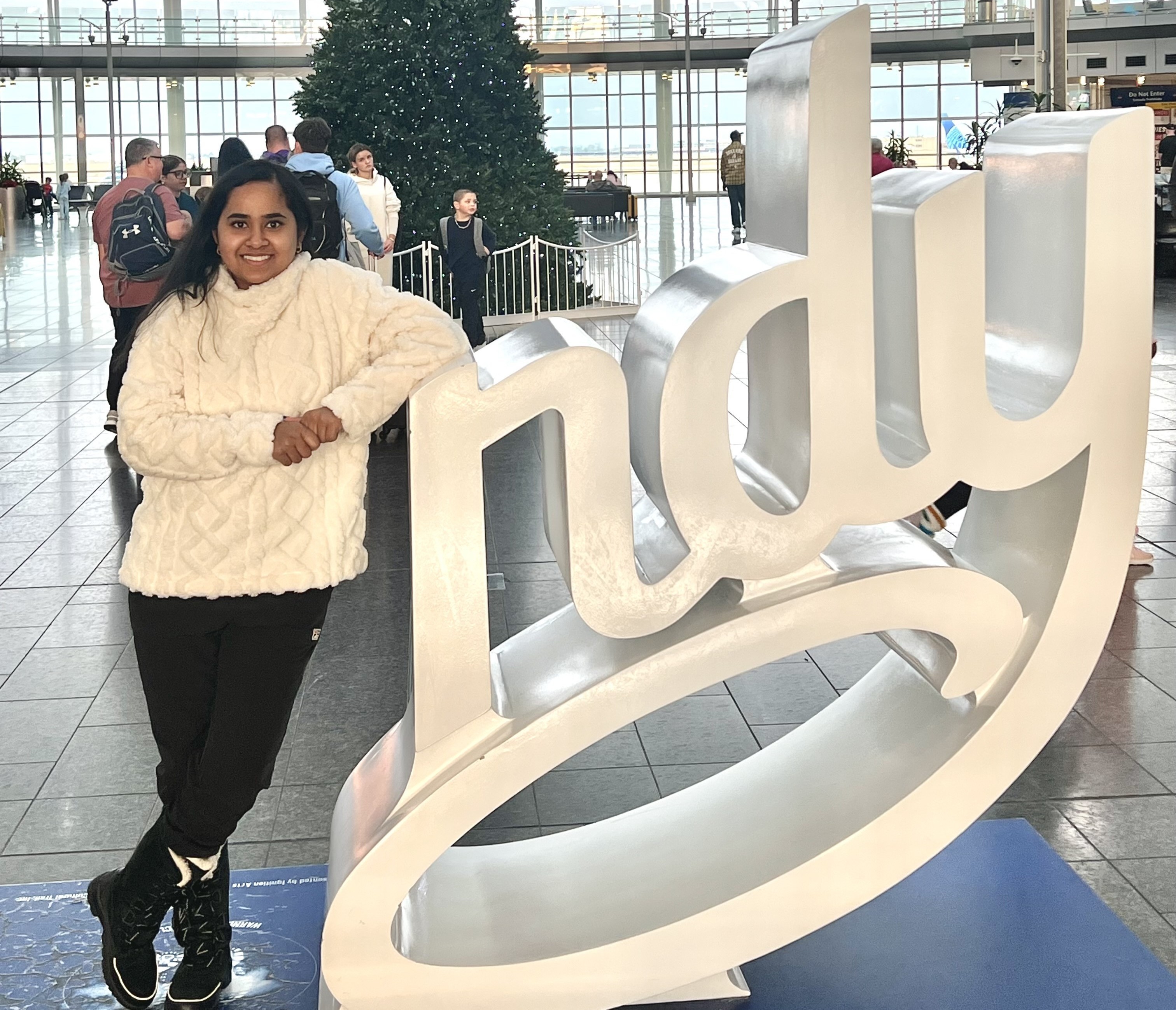Rechal Parmar by Indy Sculpture Rechal Parmar, wearing a white winter sweater, black pants, and black winter boots leans against a sculpture of the letters "ndy" in a large building with a Christmas tree in the background