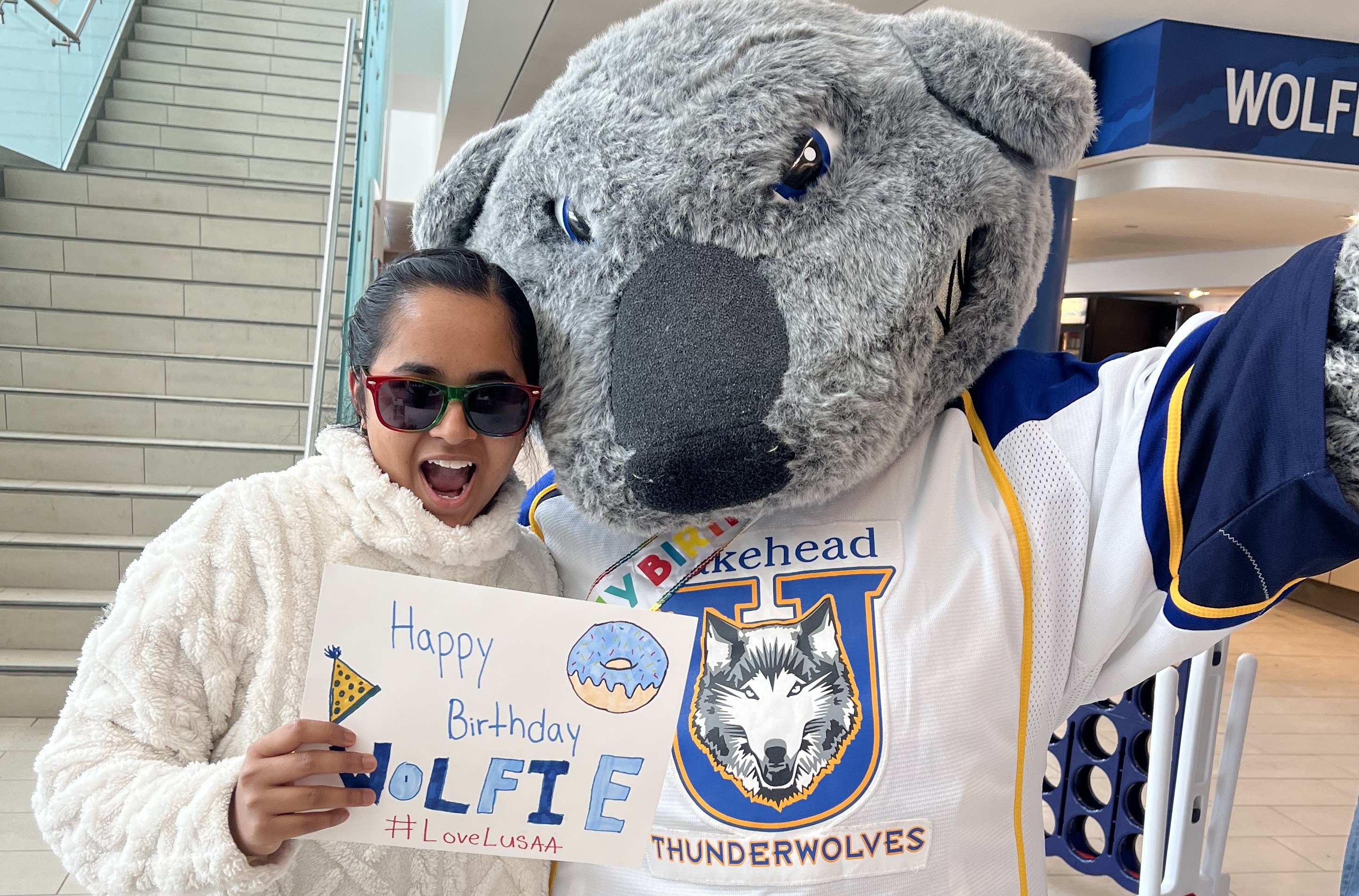 Rechal Parmar with Wolfie Rechal Parmar, wearing sunglasses and a white sweater, stands beside the Wolfie mascot and holds a sign saying "Happy Birthday Wolfie #LoveLUSAA"