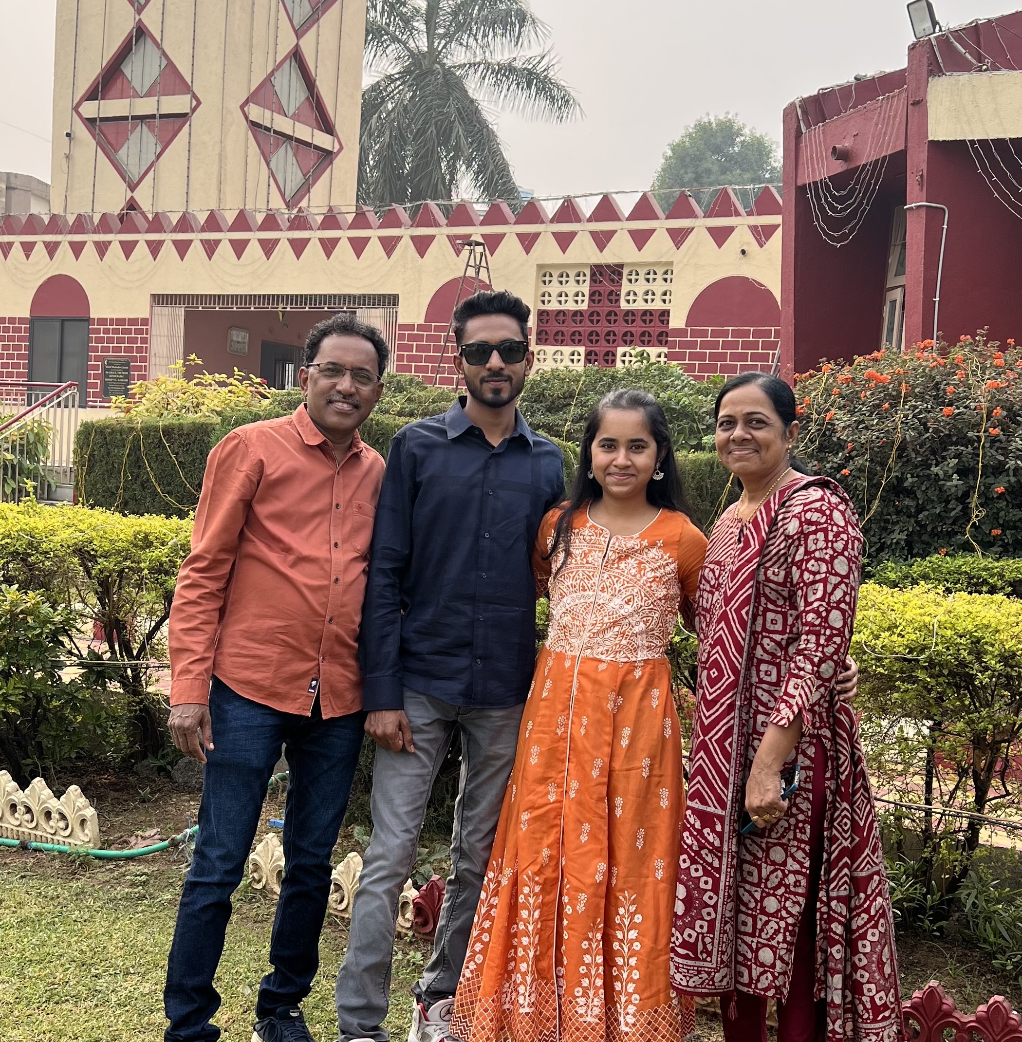 Rechal Parmar, wearing a long orange-patterned dress stands in a garden with her father, brother, and mother; in the background is a large red and cream-coloured building with geometric shapes and a palm tree