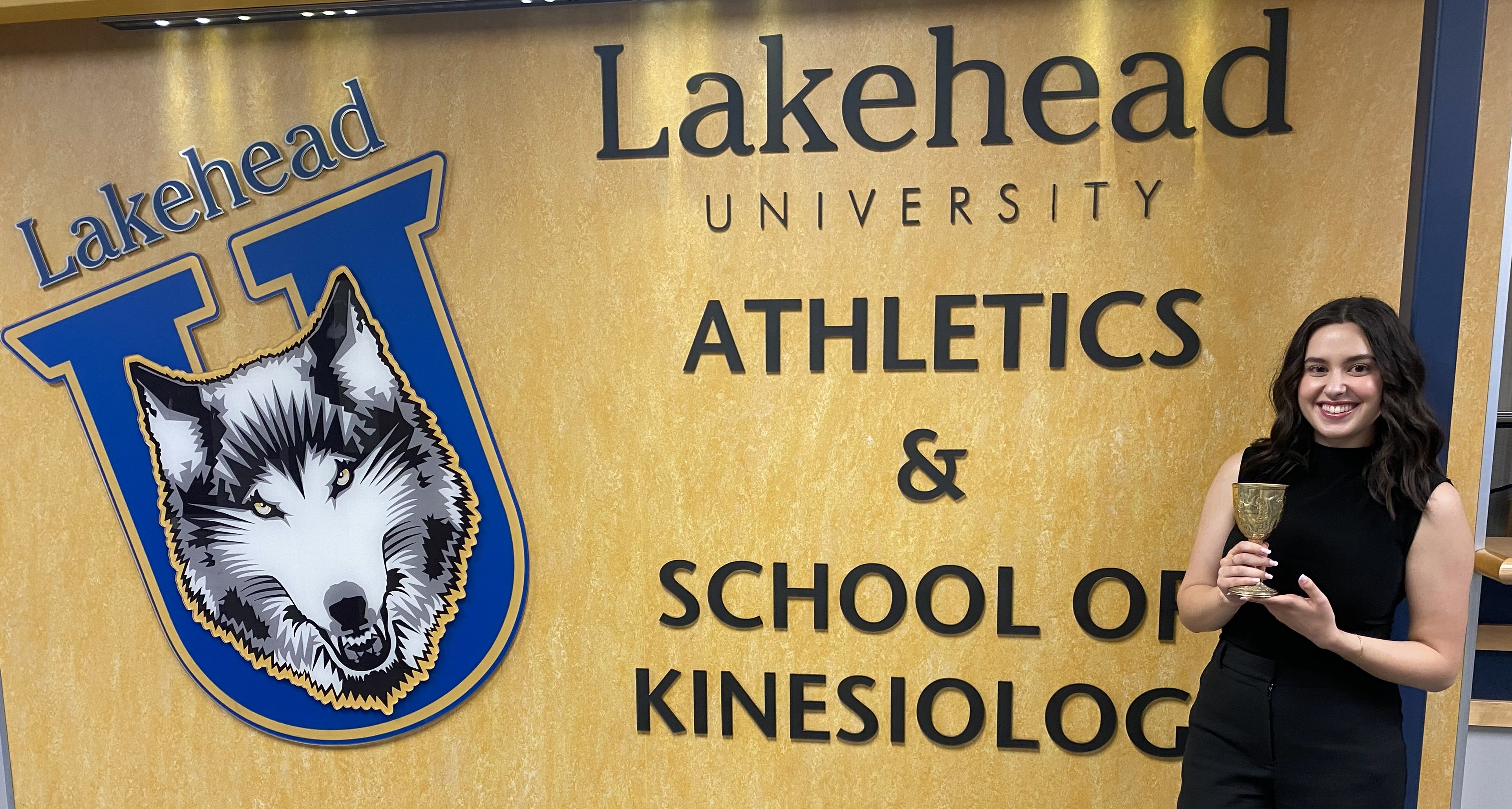 Chalice of Knowledge Madeline Fabiano, holding a gold-coloured chalice, stands in front of a wall with the Lakehead Thunderwolves logo and that bears the words Lakehead University Athletics & School of Kinesiology