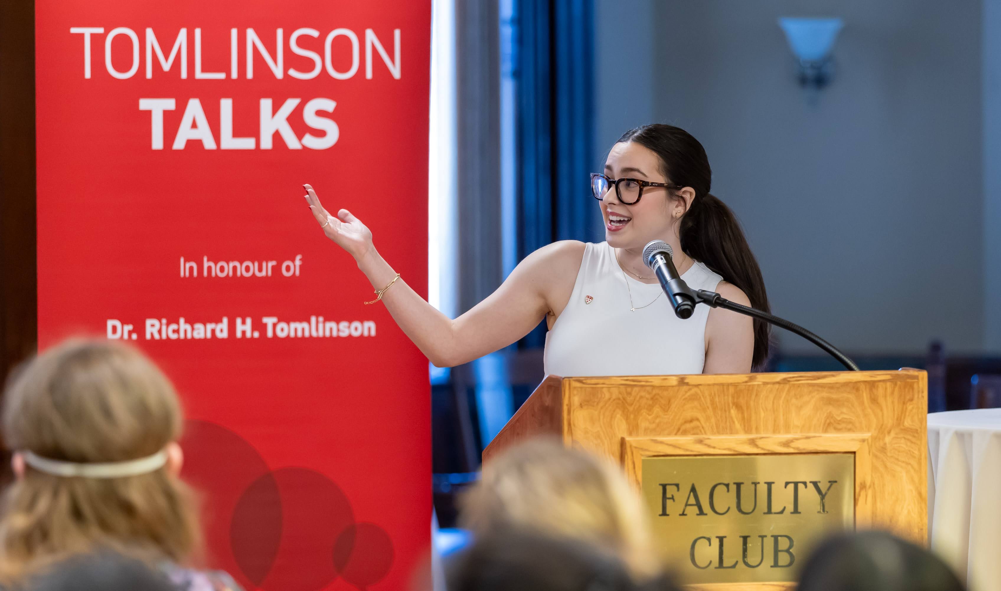 Madeline Fabiano Gives a Presentation Madeline Fabiano stands in front of a wooden podium (bearing the words "Faculty Club") with a microphone and gestures with one hand; beside the podium is a red banner with the words "Tomlinson Talks In honour of Dr. Richard H. Tomlinson
