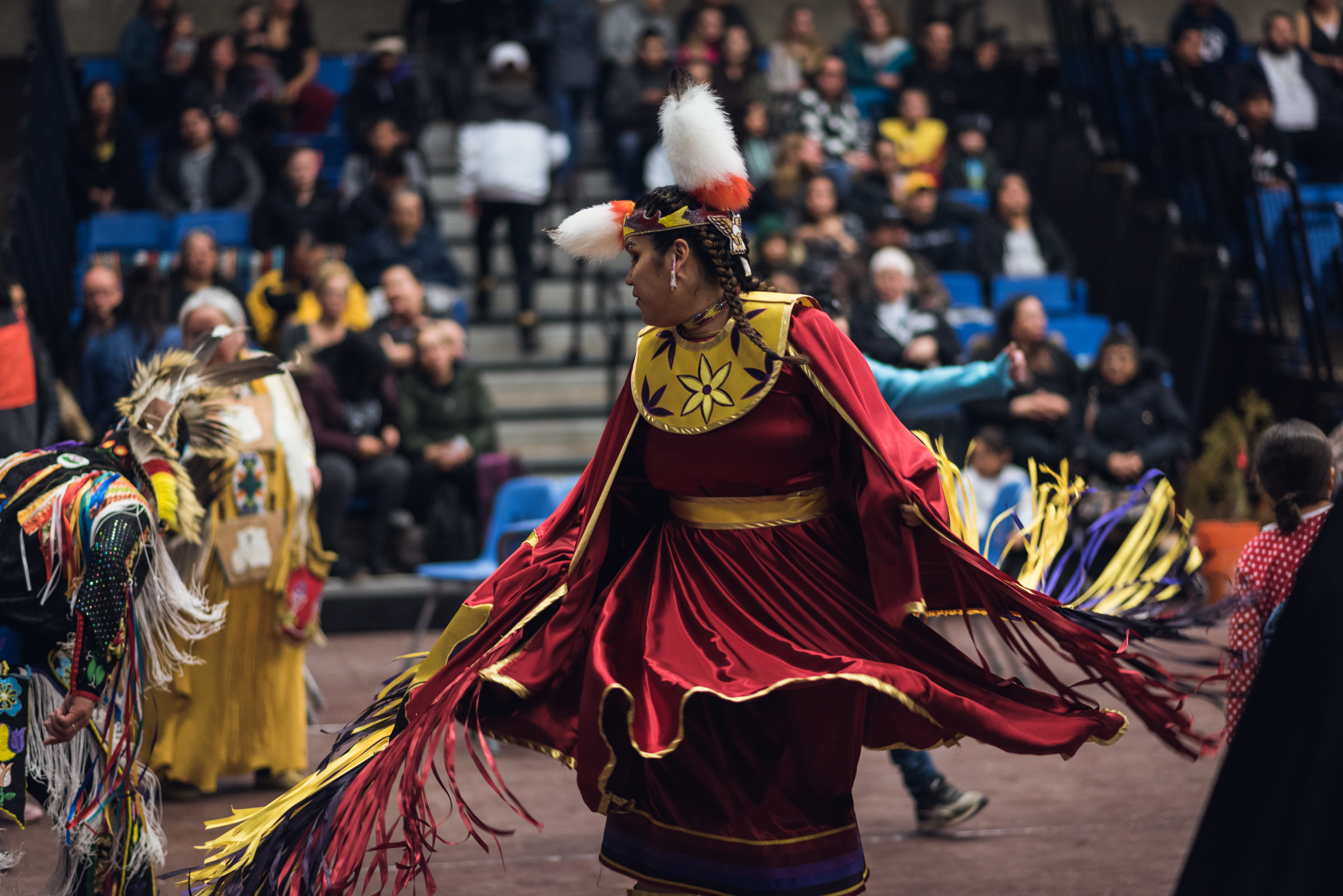 A woman with braided hair wearing a red-and-yellow dress dances in a Powwow in front of an audience seated in bleachers