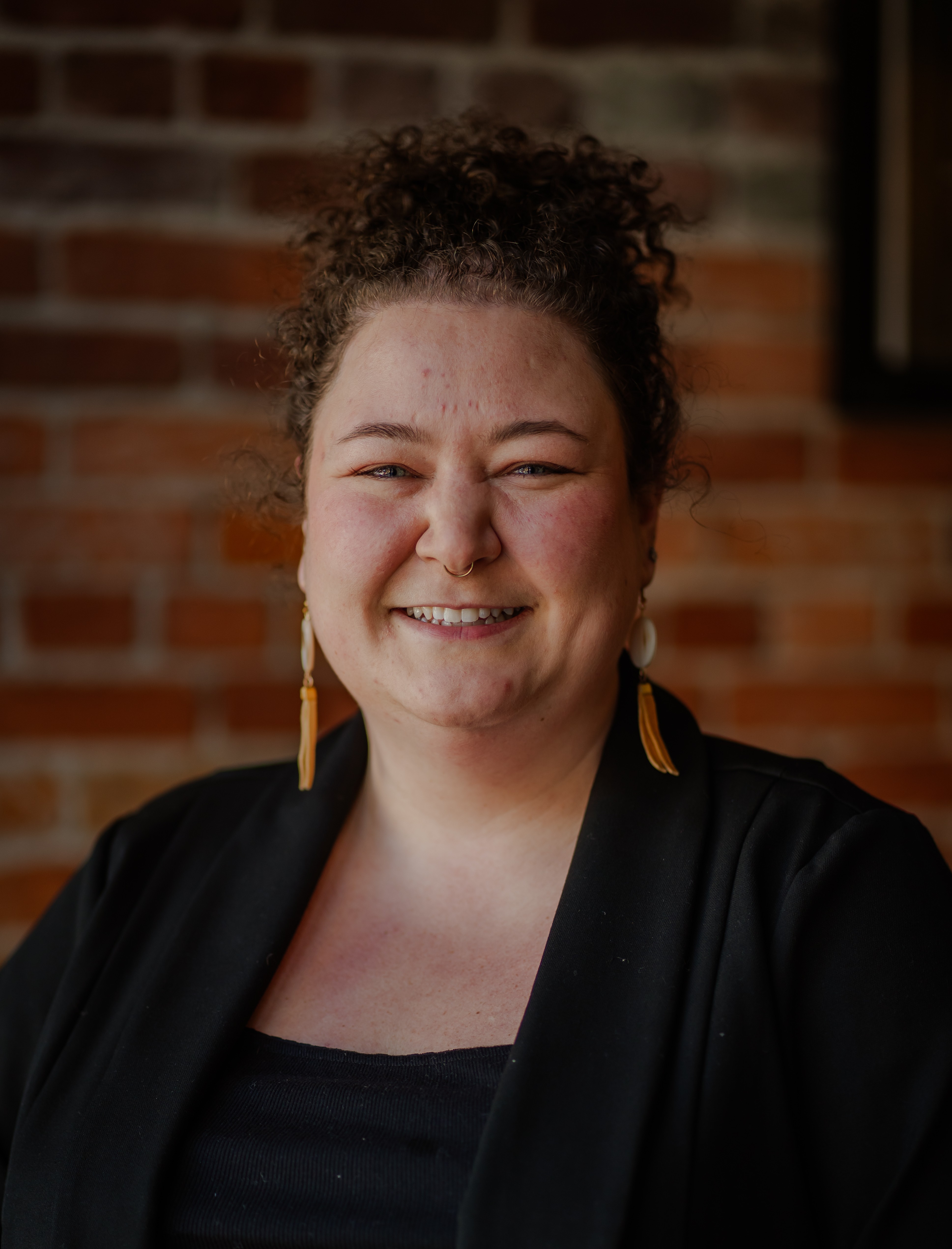 A head-and-shoulders photo of Teagan Neufeld smiling and wearing a black top and black blazer and standing in front of a brick wall