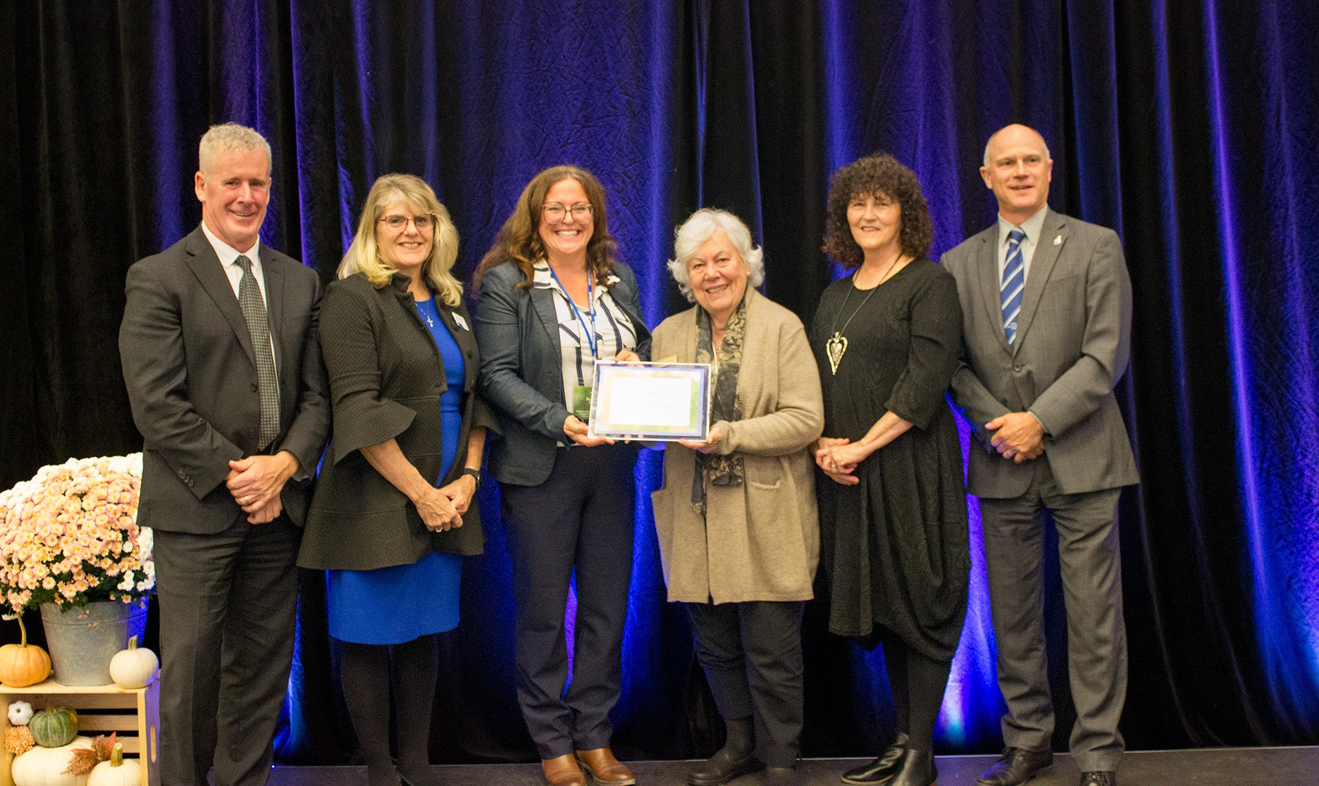 2026 Simcoe County Age-Friendly Award Ceremony A group of people stand on a stage with a blue curtain behind them; two women in the centre hold a plaque