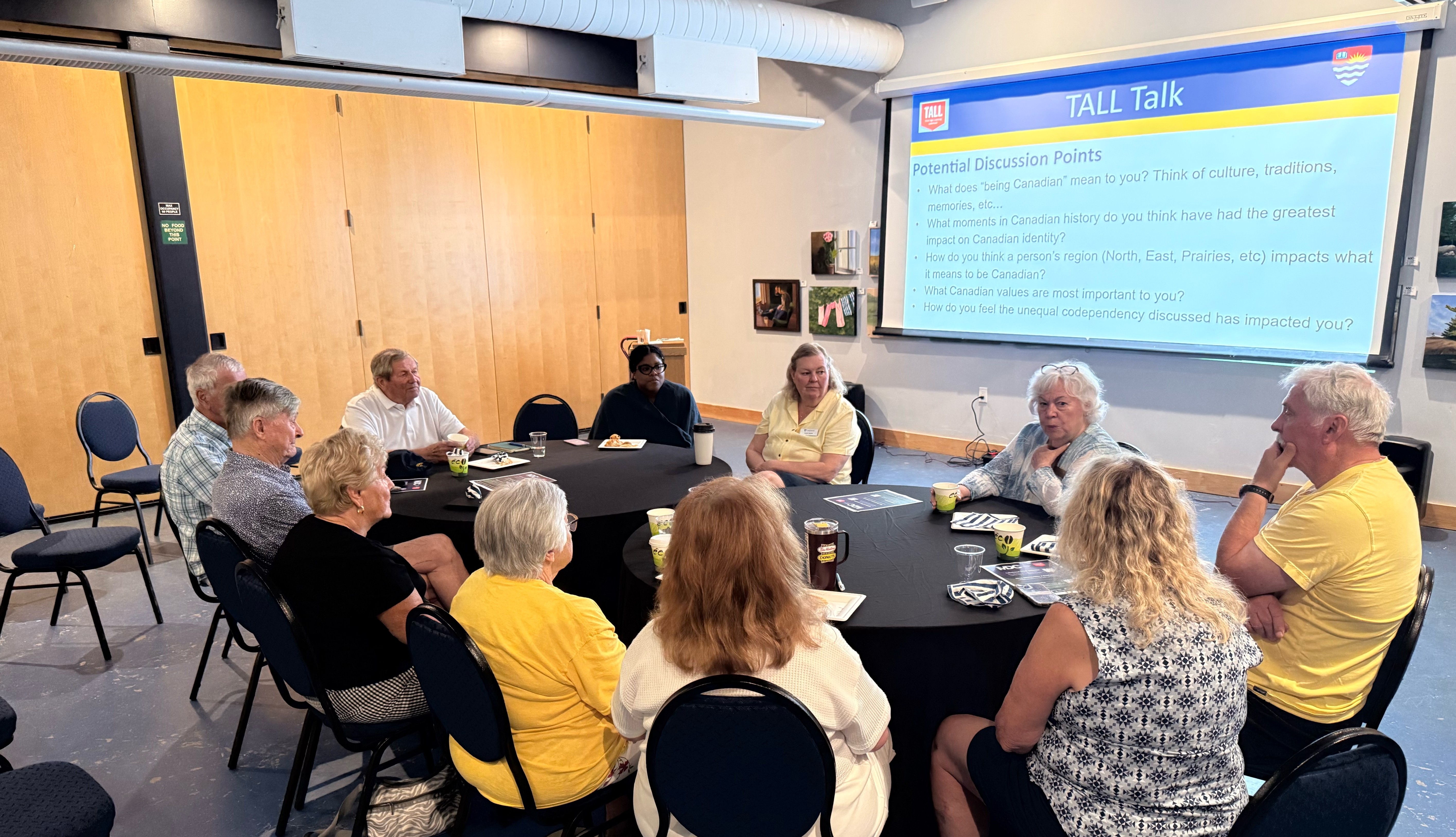 TALL Committee Members A group of older adults, and one young person, sit at two tables in a room with cabinets and a large display screen
