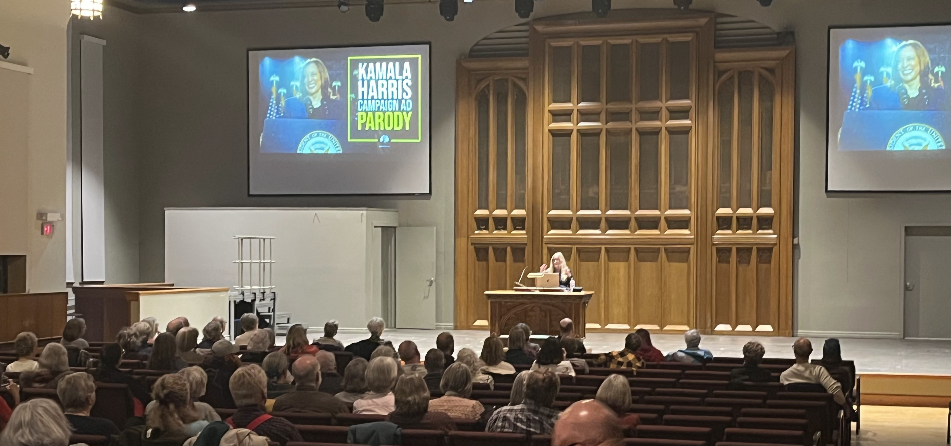 TALL Disinformation Lecture An audience in a church listen to a woman speak on stage; there are two large projection screens on the wall