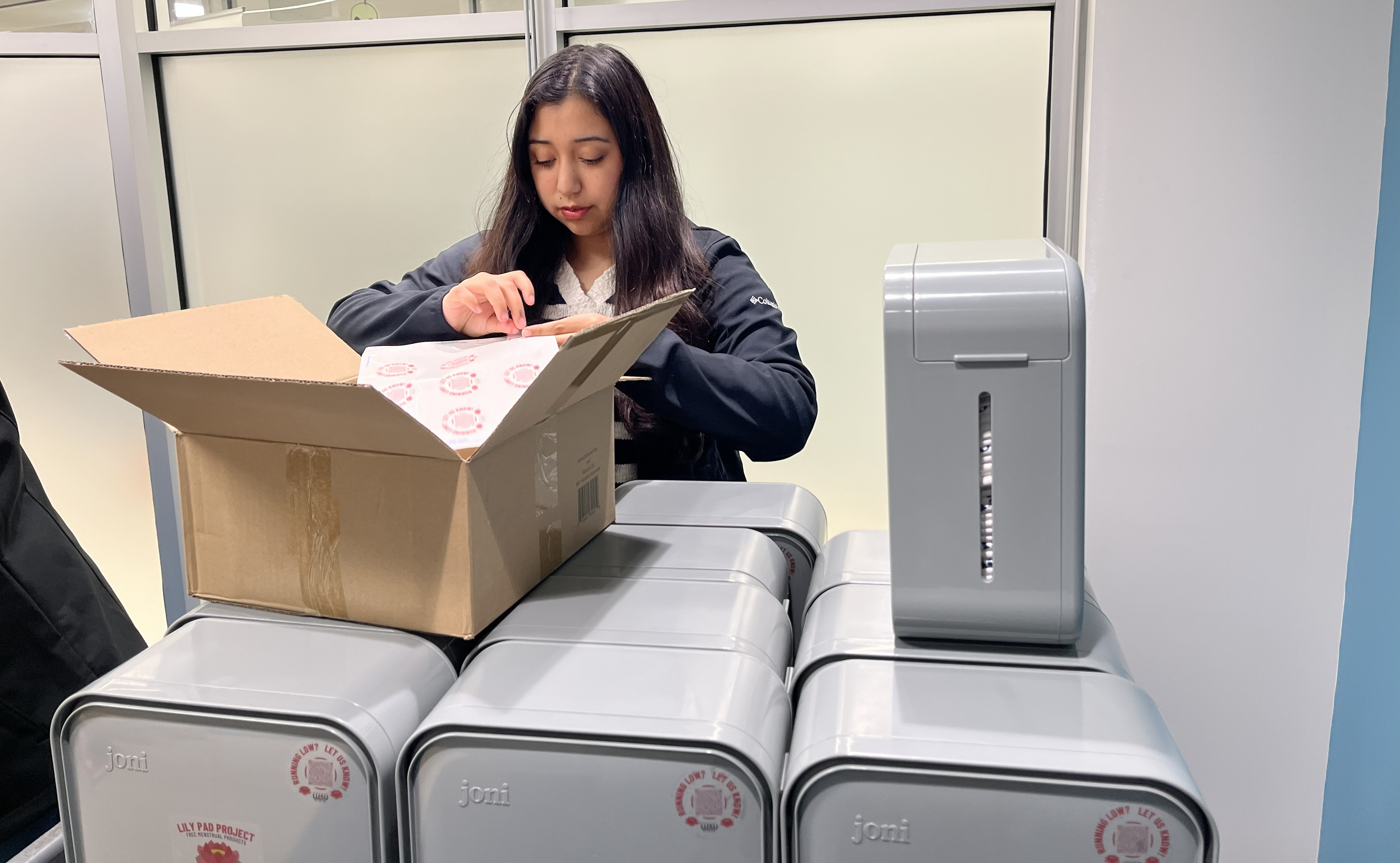 Aliss Chavarri Stocks Lily Pad Dispensers Aliss Chavarri stands in LUSU's office peeling a label off a sheet to apply to a Lily Pad dispenser