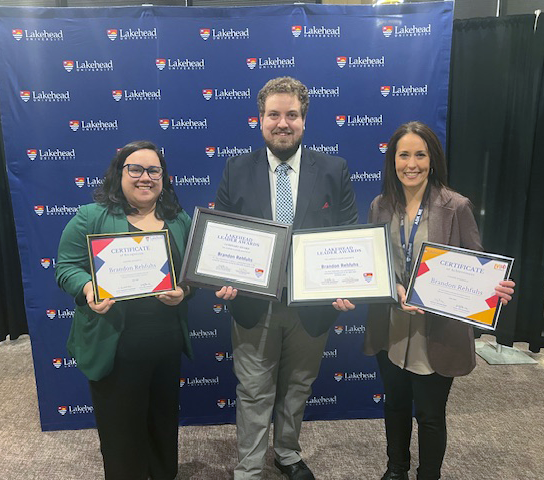 Brandon Rehfuhs holds two framed students awards while flanked by Paula Jean Broderick and Andrea Tarsitano