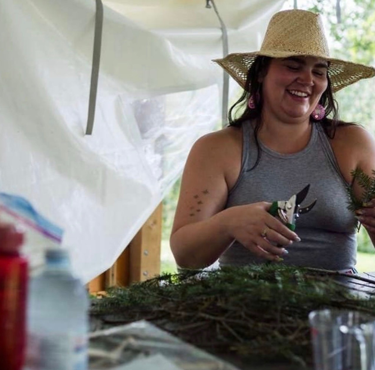 Céline Wick Processes Medicines at a Medicine Camp Céline Wick smiles as she holds a pair of clippers in one hand and a piece of a spruce tree bough in her other hand; a pile of tree boughs sit on a table in front of her