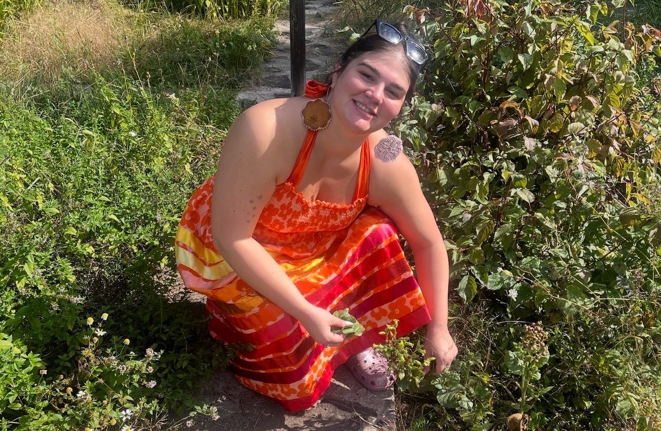 Céline Wick in Gitigaan Medicine Garden Céline Wick, wearing an orange dress and beaded earrings, picks leaves from plants in a garden on a sunny day